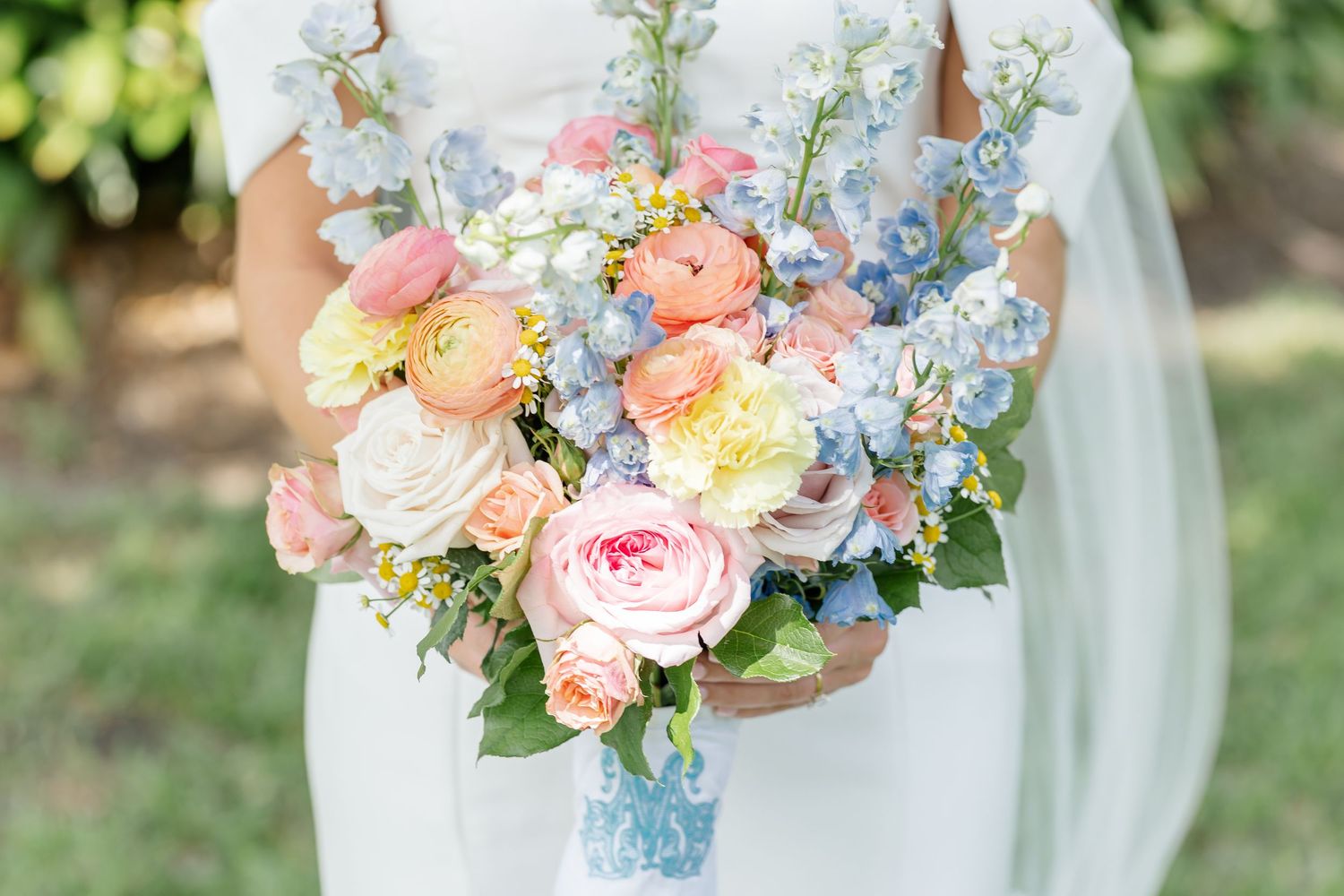 Pastel spring wedding bouquet featuring pink roses, peach ranunculus, and blue delphinium.