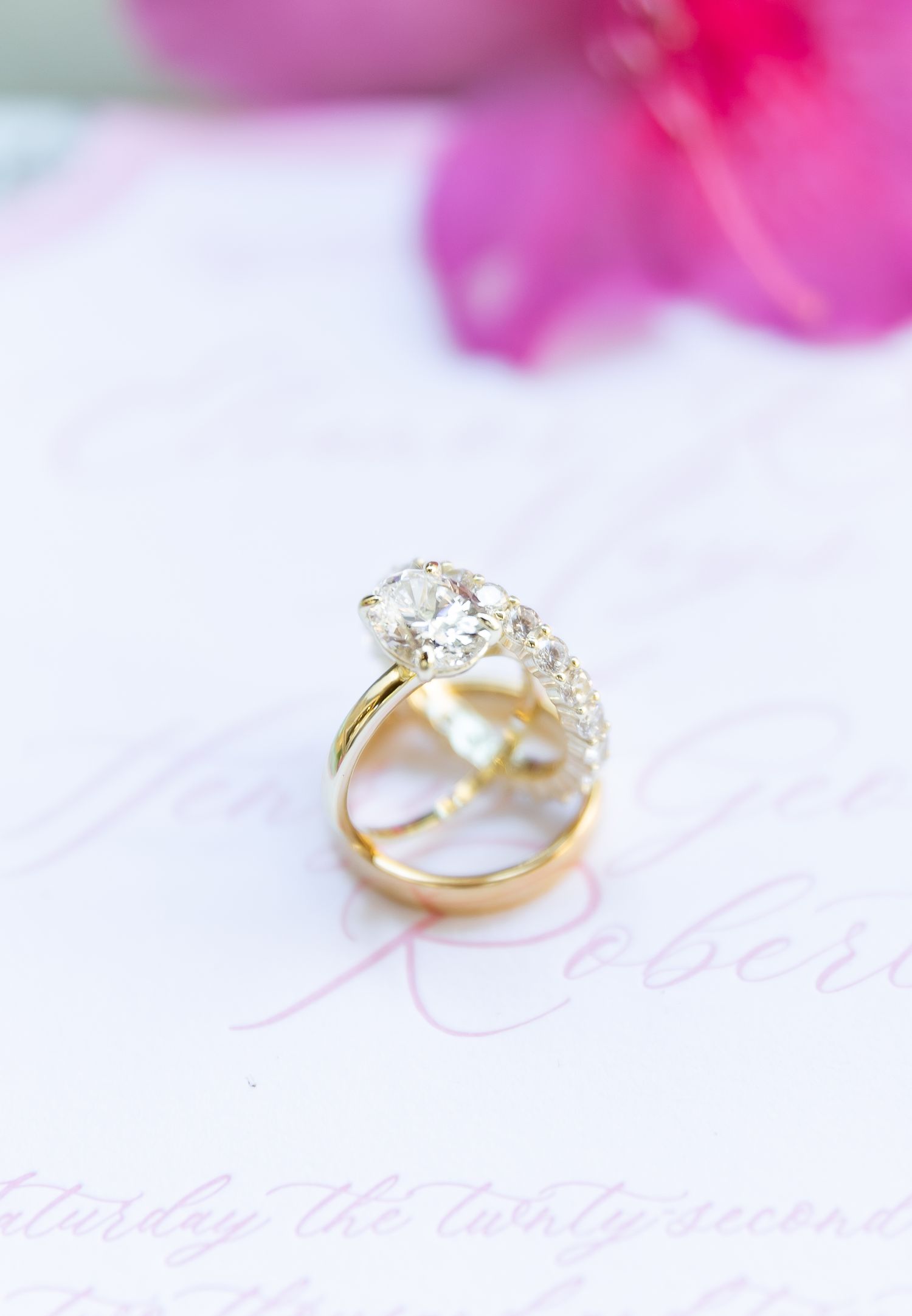 Gold diamond engagement ring photographed with soft pink flower bokeh background on white surface.