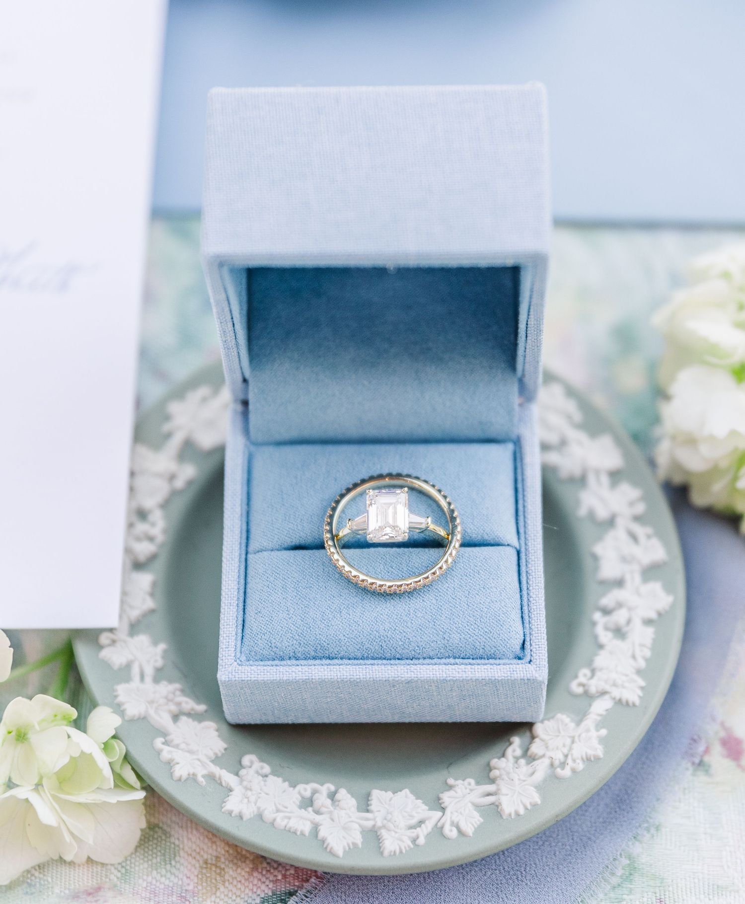 A close-up of a diamond engagement ring in a light blue velvet jewelry box with delicate floral decor in the background.