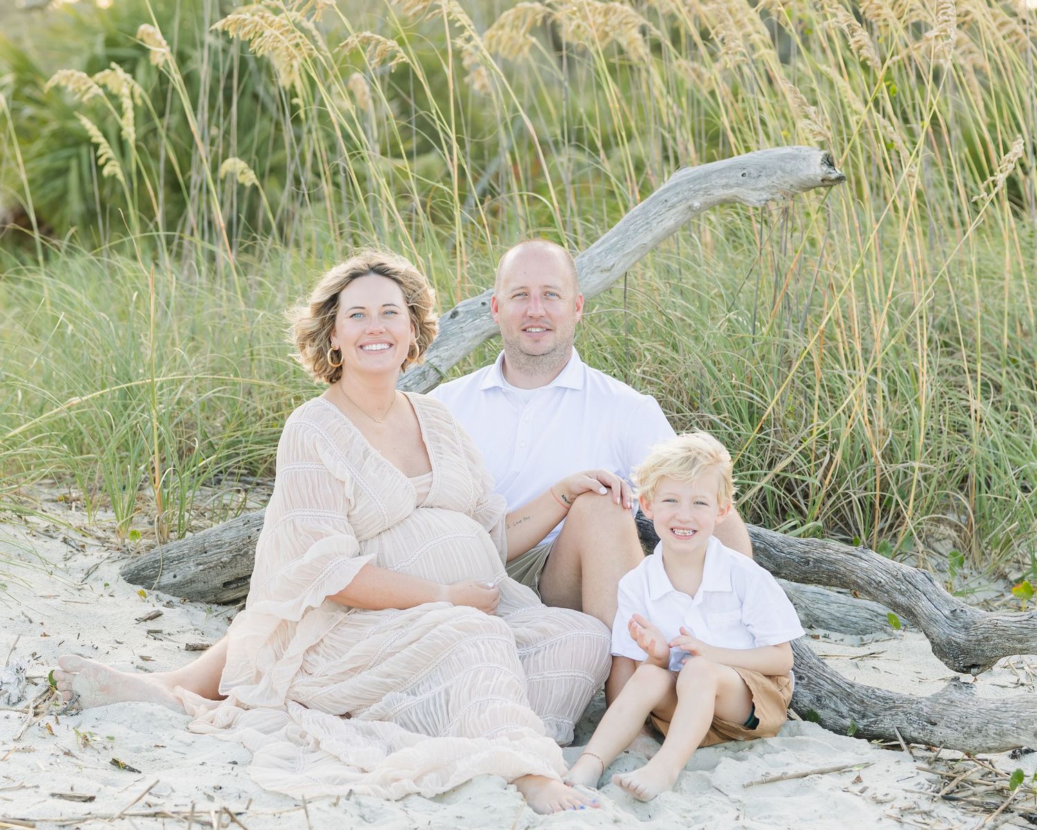 Family sits together on driftwood at beach surrounded by sea grass at sunset.