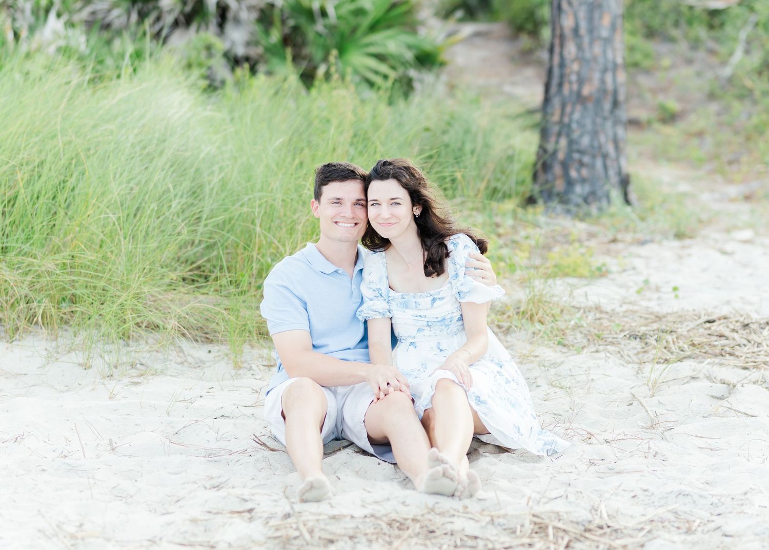 A couple dressed in light blue and white sits together on a sandy beach with tall grass and trees in the background.