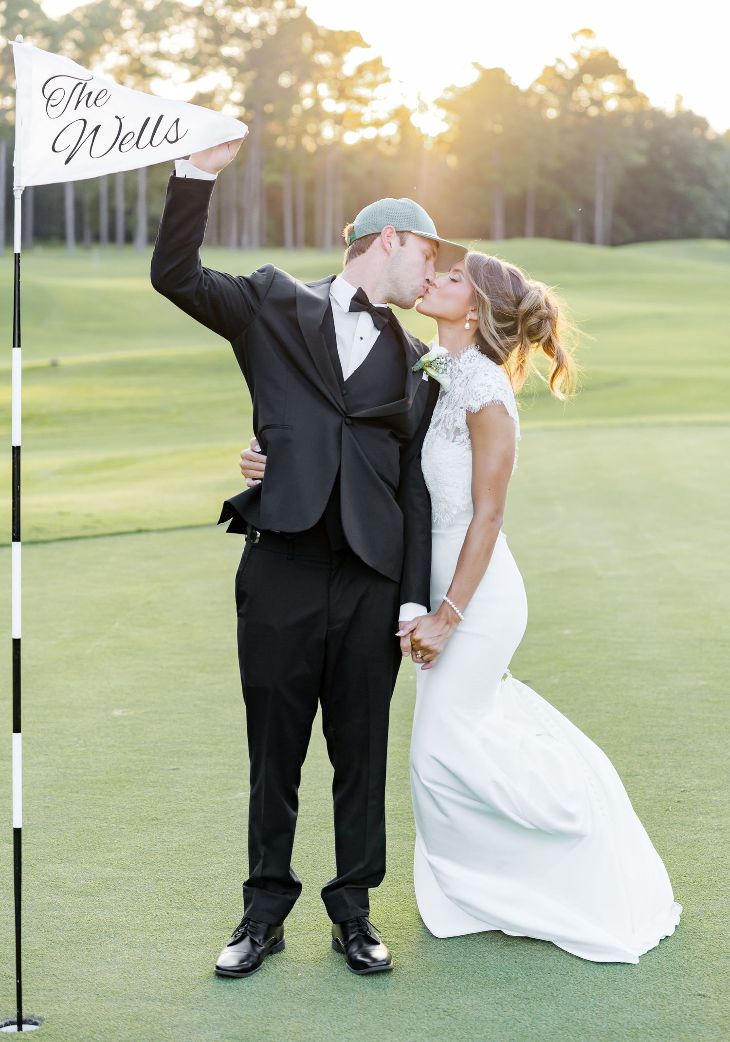 Newlywed couple kissing on golf course at sunset, groom in tuxedo holding flag reading The Wells, bride in fitted white gown.