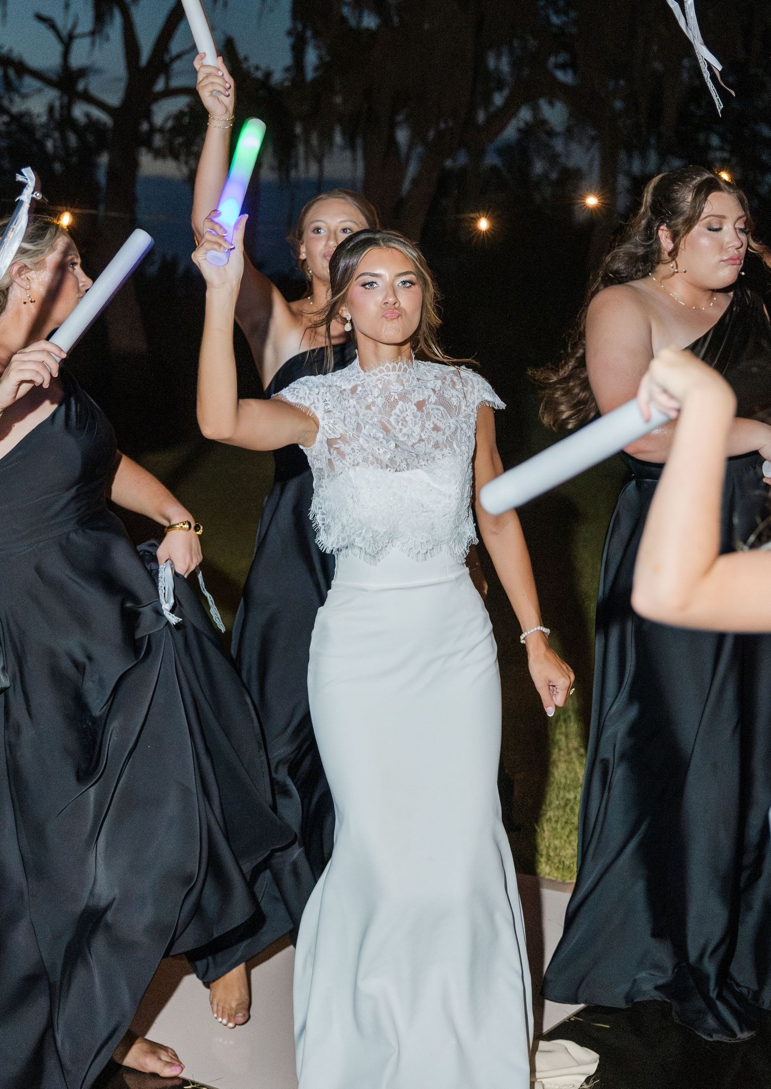 Bride in lace gown dances with bridesmaids holding colorful glow sticks at evening wedding celebration.