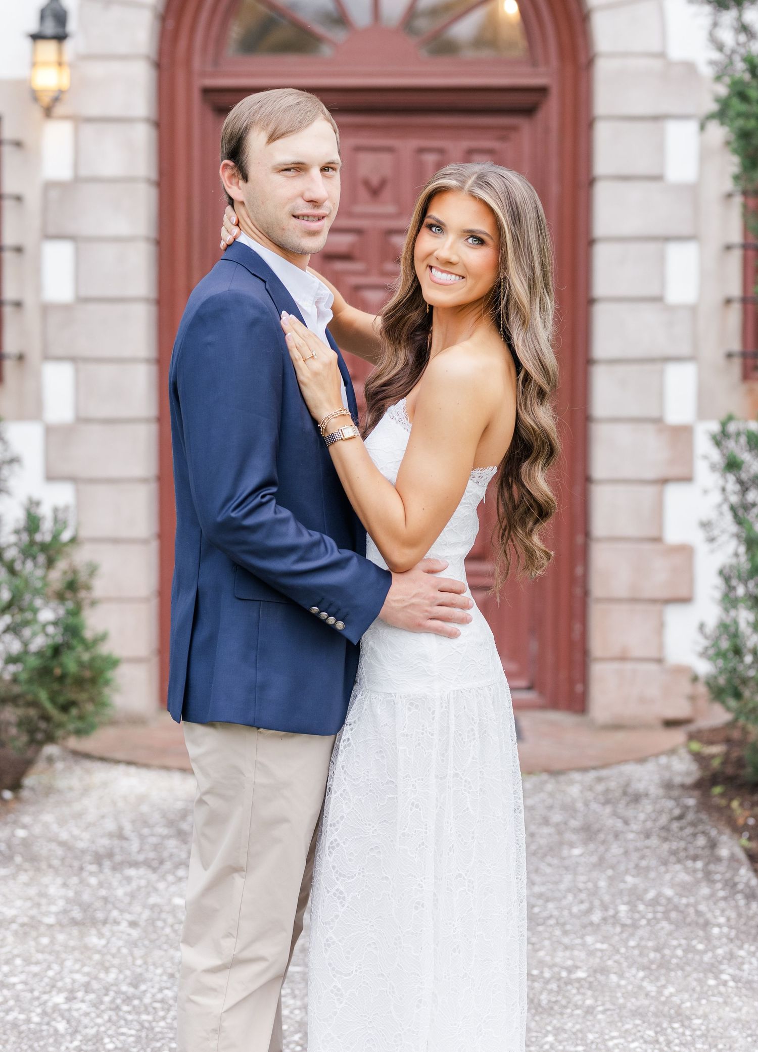 Engaged couple embracing in front of red door, man in navy suit and woman in white lace dress posing.