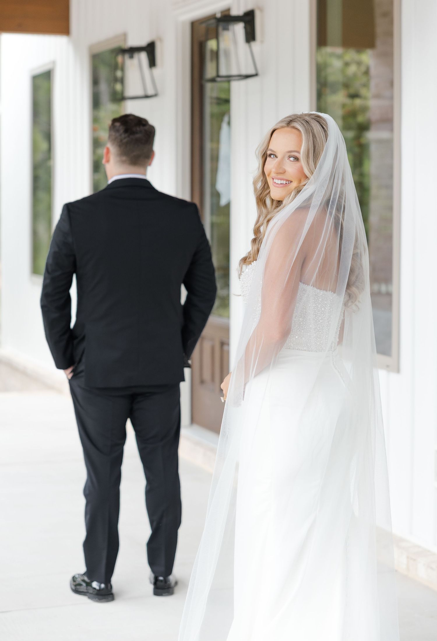 Bride in white gown and veil smiling over shoulder at groom in black suit during first look.