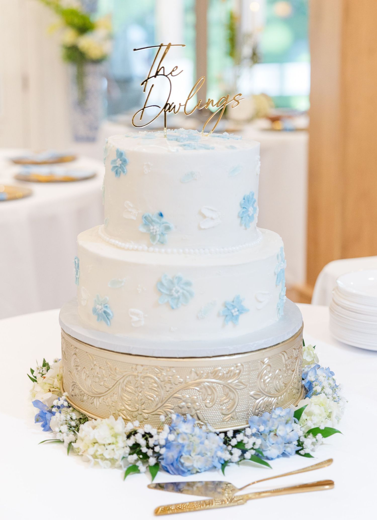 Three-tier white wedding cake with blue floral details and gold 'The Dawlings' topper on ornate stand.