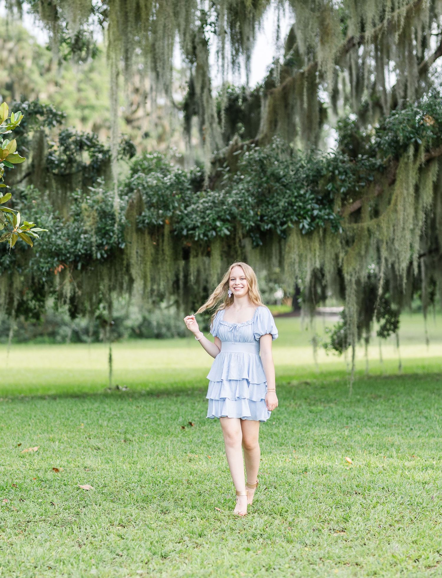 Woman in pale blue dress walks through grassy area under Spanish moss covered trees.