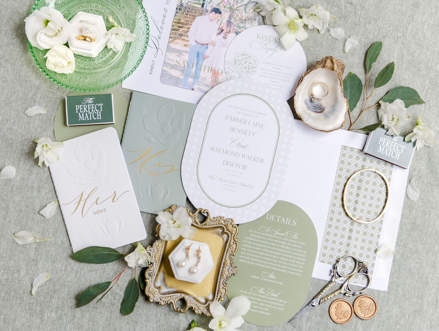 Wedding flat lay featuring sage green stationery, white roses, gold rings, earrings, and elegant invitation suite.