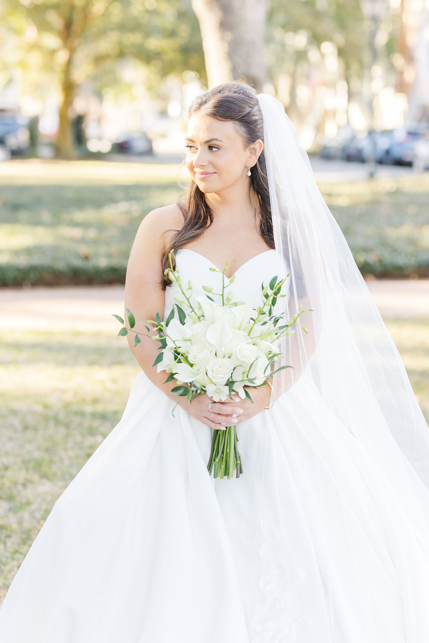 Bride in strapless white ball gown with cathedral veil holds white floral bouquet in sunny park setting.
