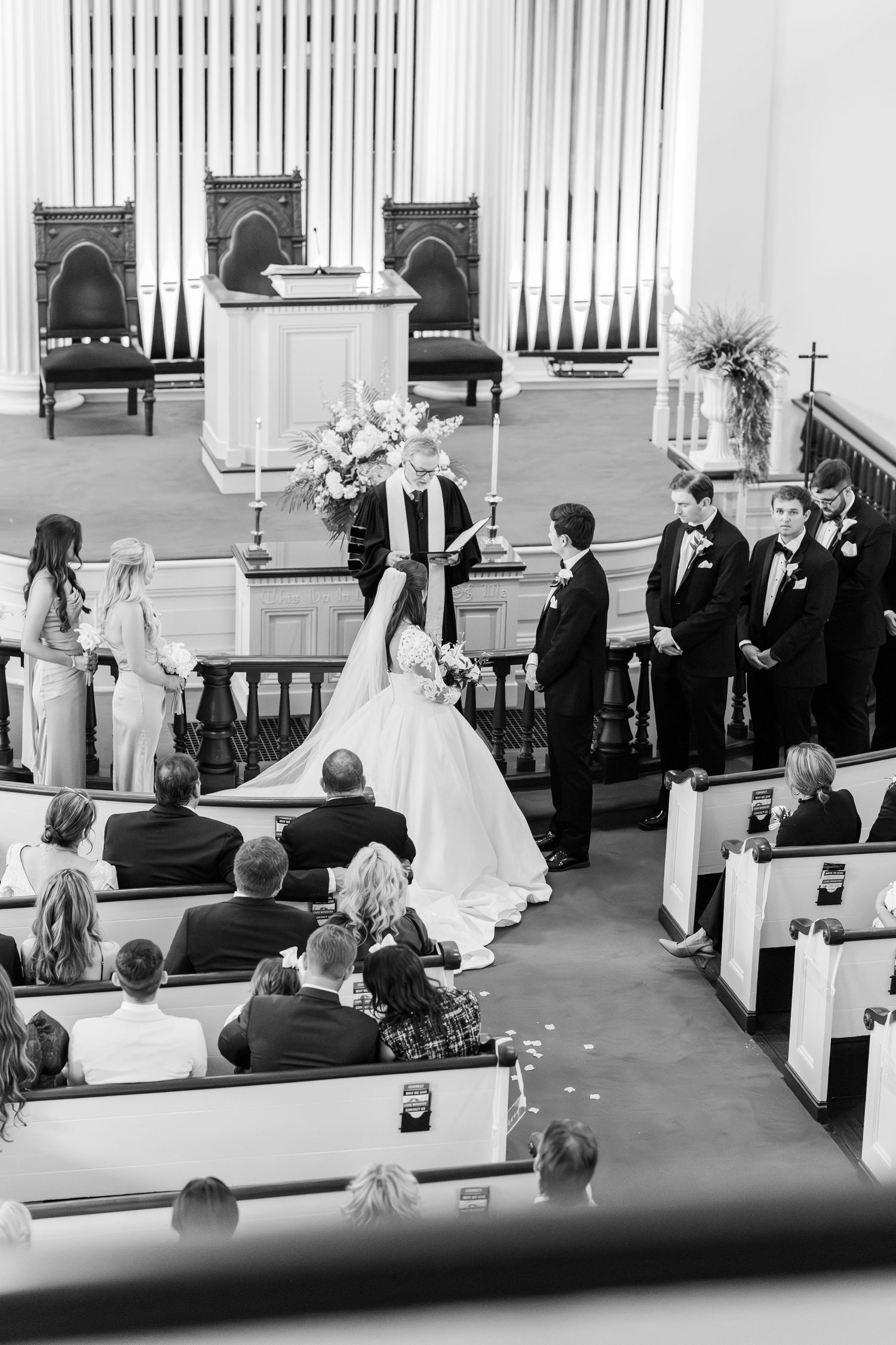 Black and white photo of bride and groom exchanging vows at church altar with wedding party.