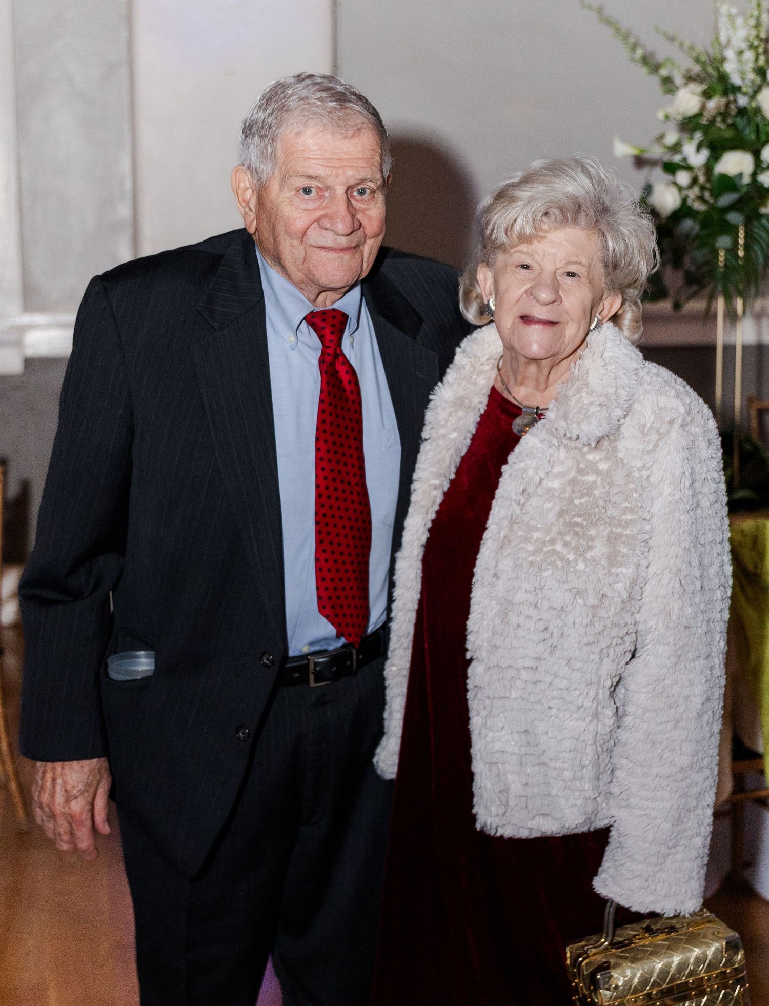 Elderly couple posing together at wedding reception, man in navy suit with red tie.