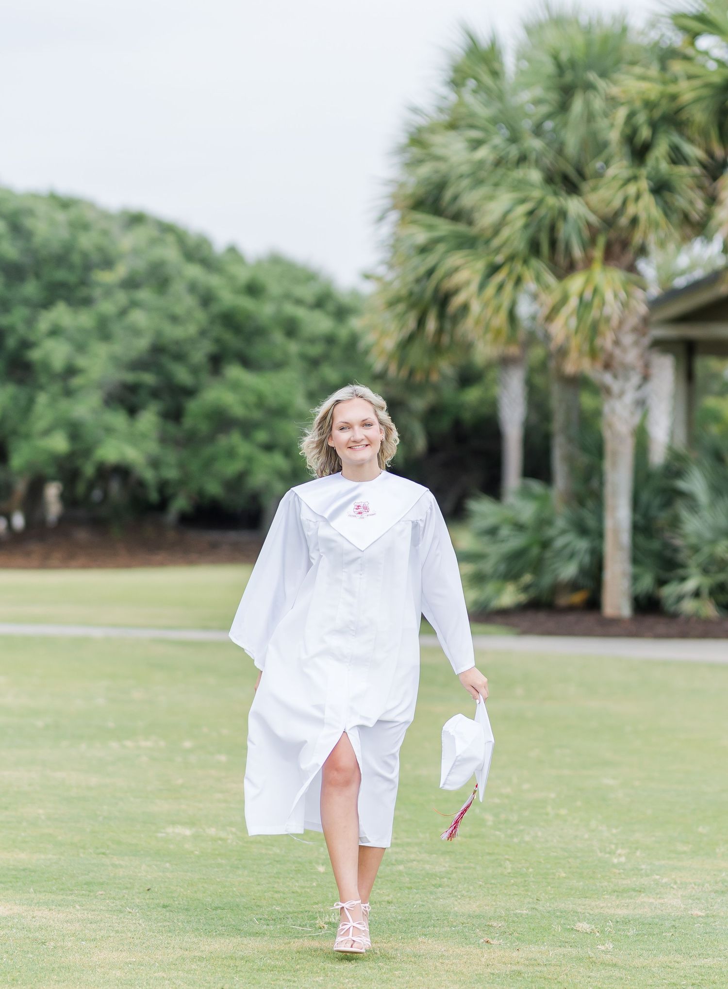 Someone in a flowing white graduation gown walks across a grassy lawn with palm trees in the background.