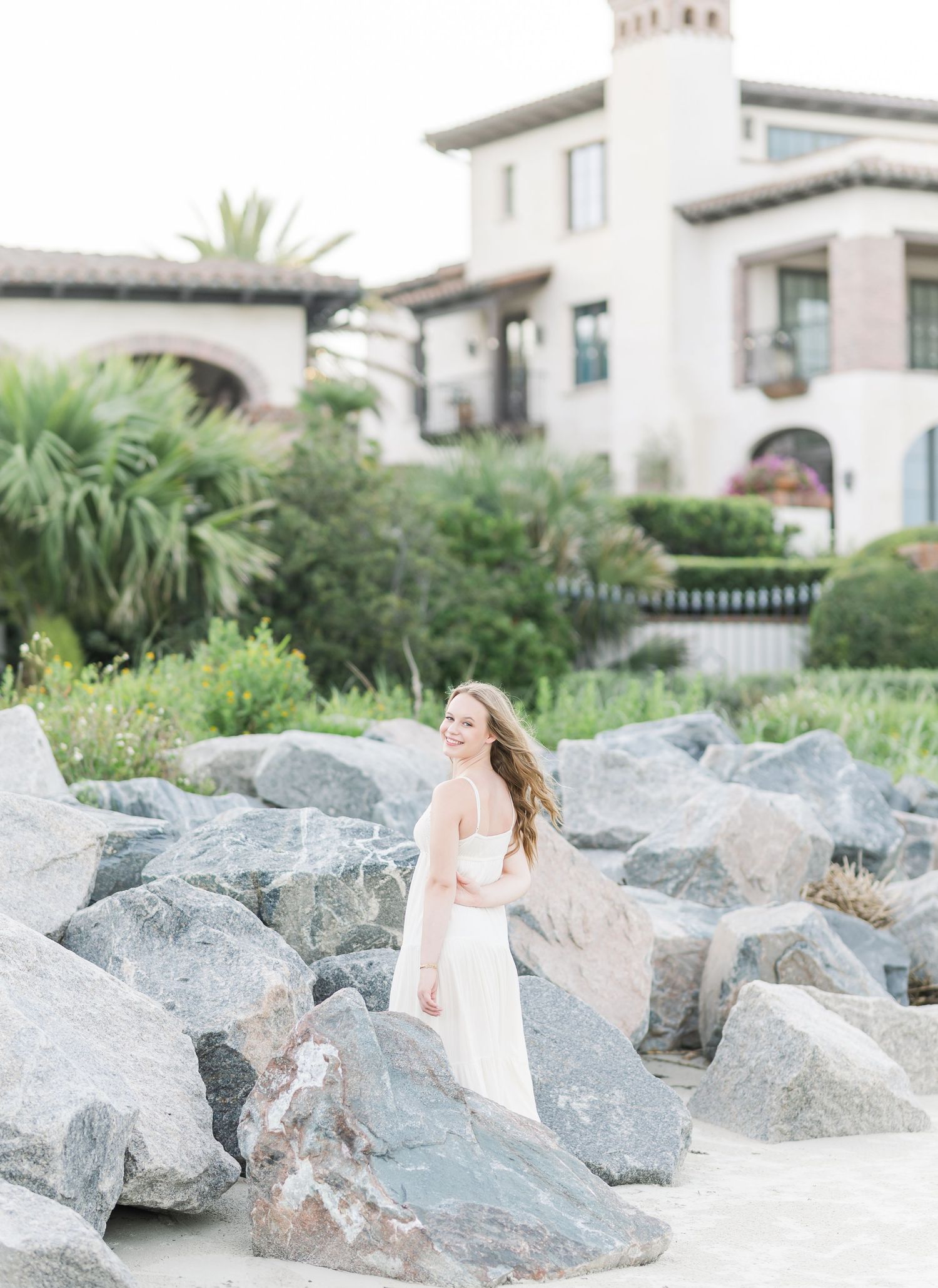 Woman in a white dress walking among large coastal rocks with beachfront houses in the background.