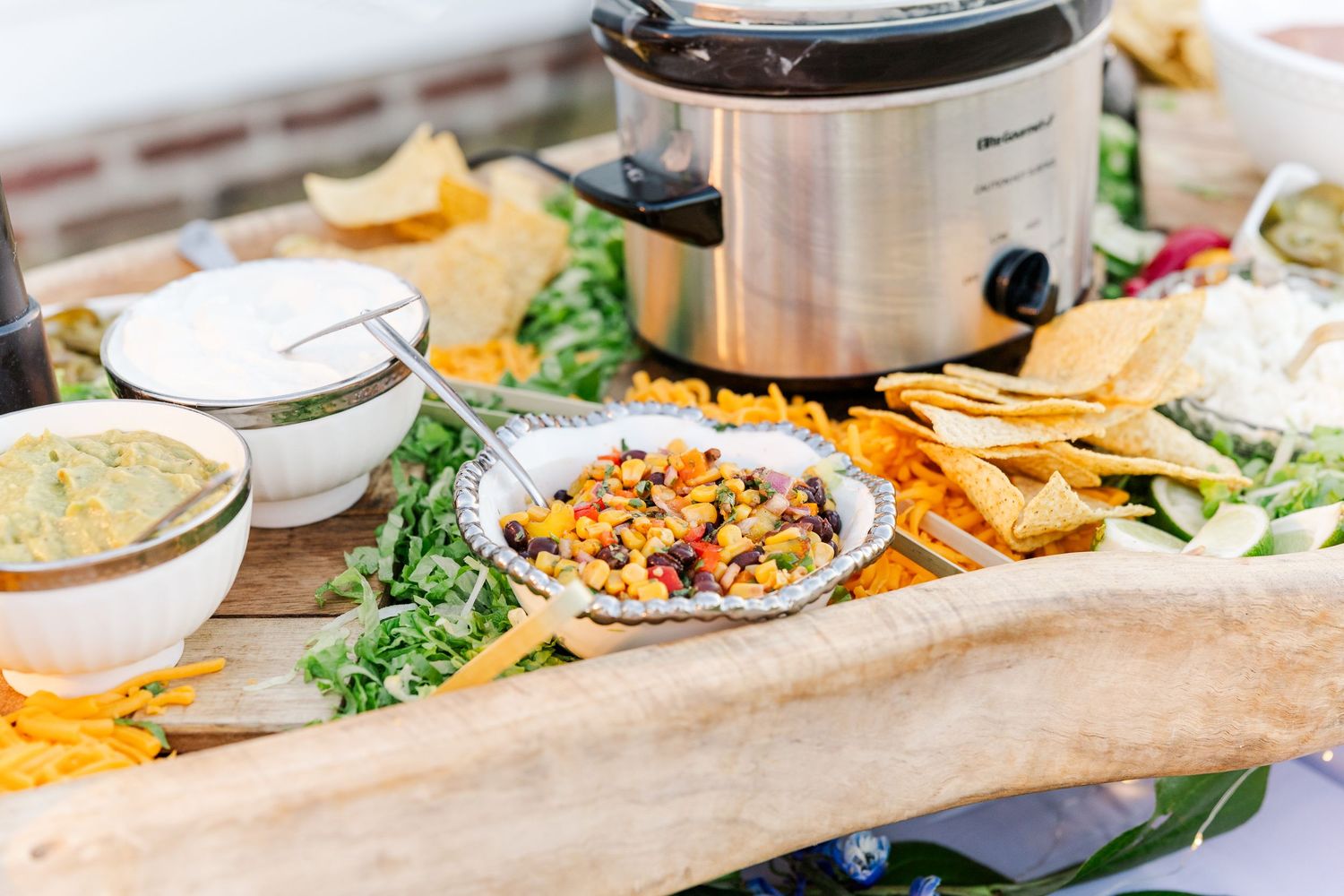 Close-up of a colorful taco bar grazing board with nachos, salsa, guacamole, and a slow cooker.
