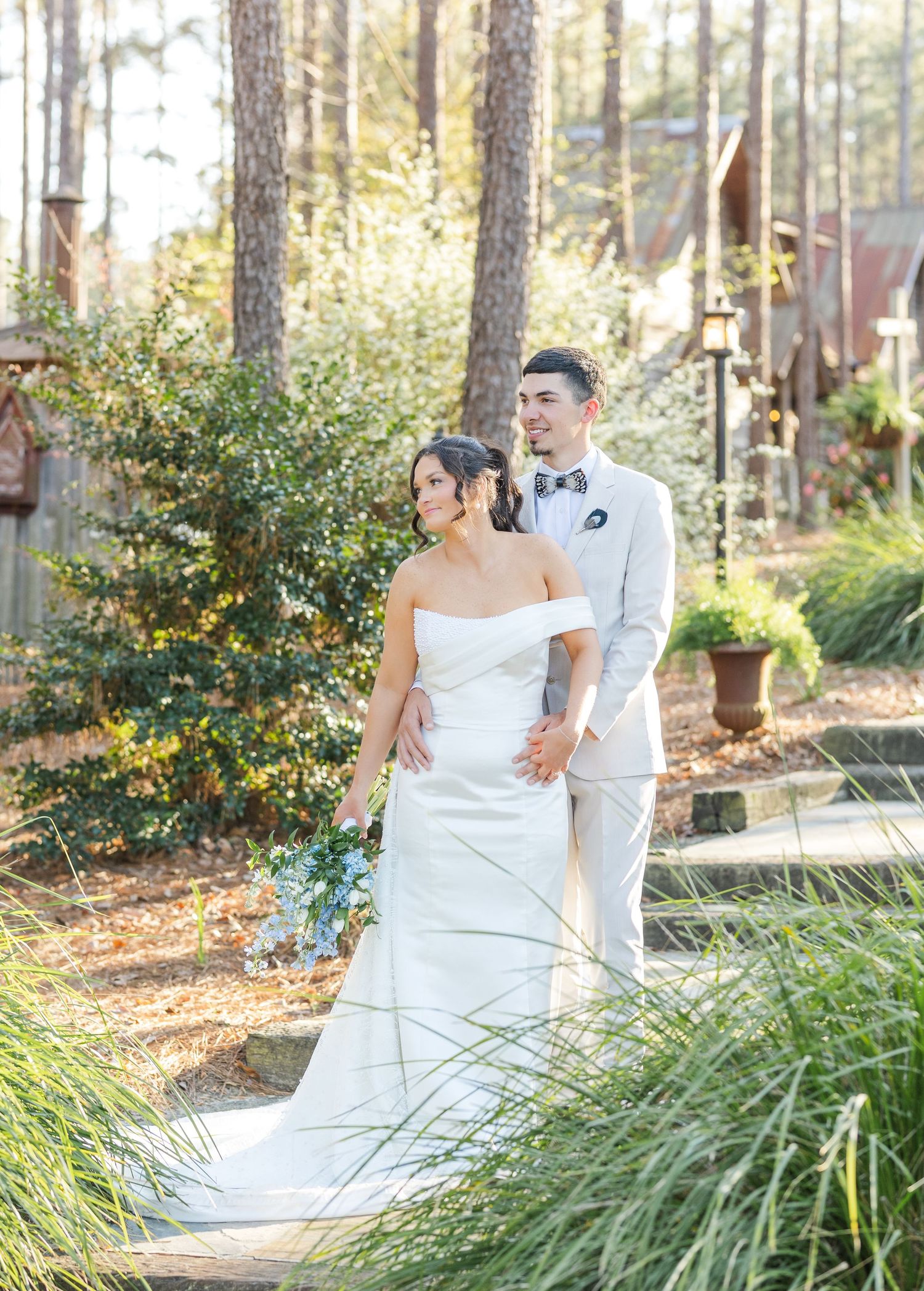 Bride in off-shoulder white gown and groom in tan suit holding hands in lush garden setting.