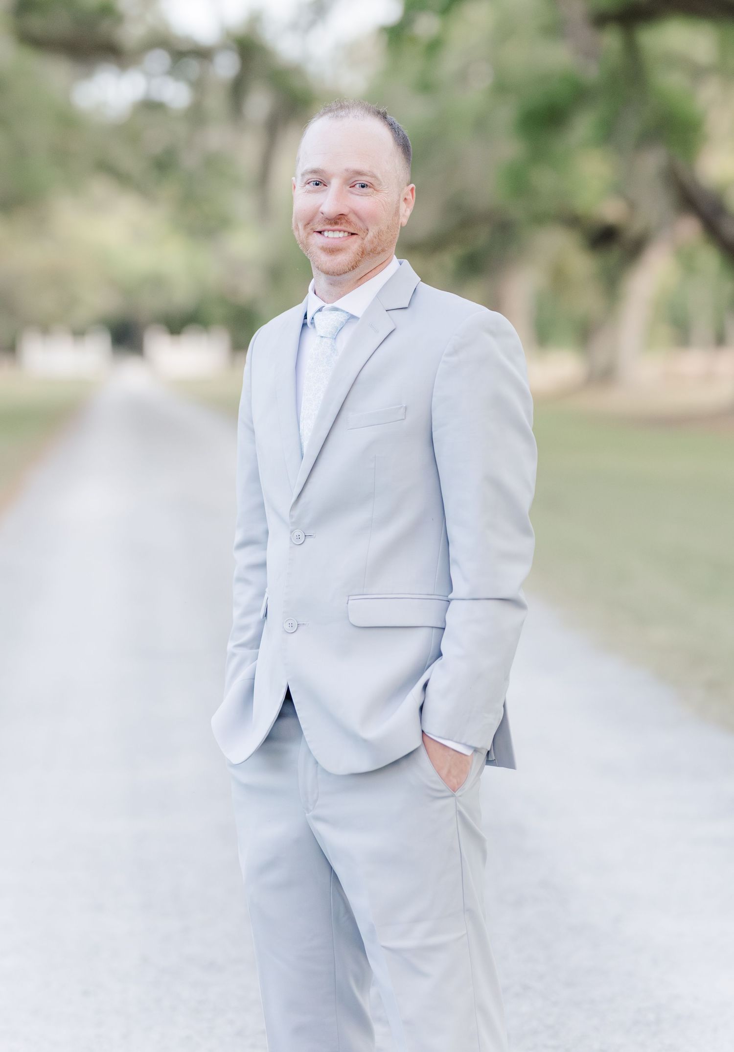Smiling groom in light gray suit standing on tree-lined path at outdoor wedding venue.