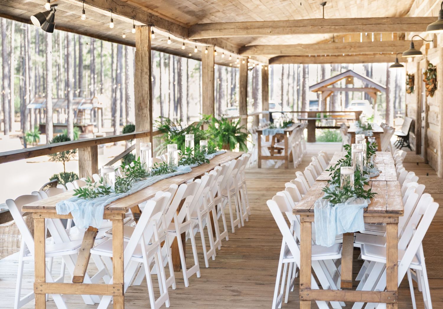 Elegant barn wedding reception with long farm tables, white chairs, and lush greenery centerpieces.