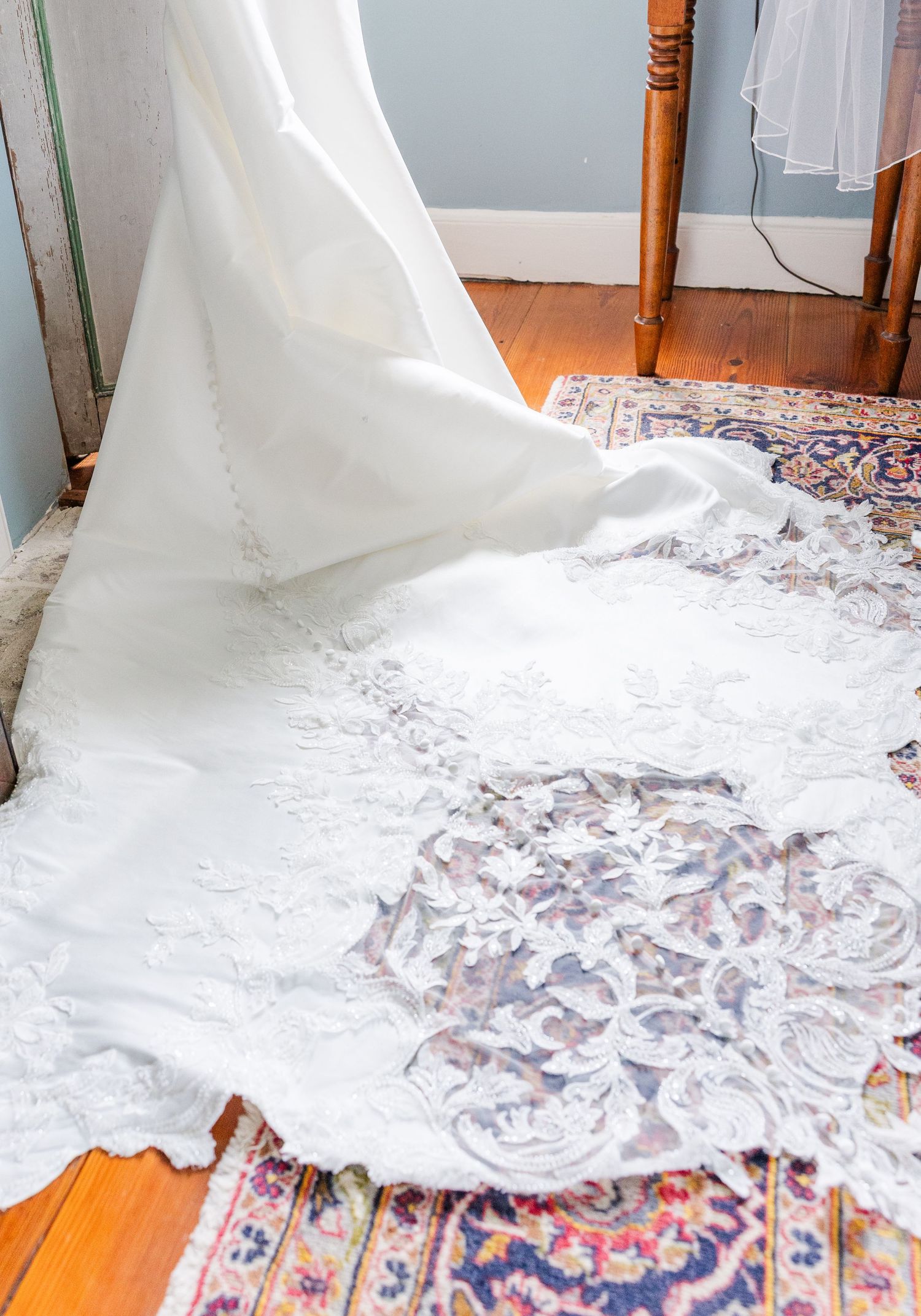 Elegant white wedding dress with lace train spread on a vintage Persian rug in a rustic room.