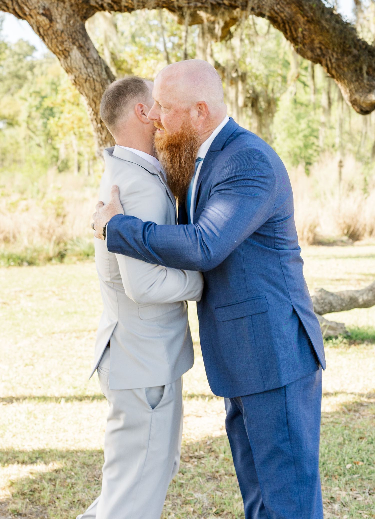 Two grooms sharing a kiss under a tree, one in a gray suit and one in a navy blue suit.