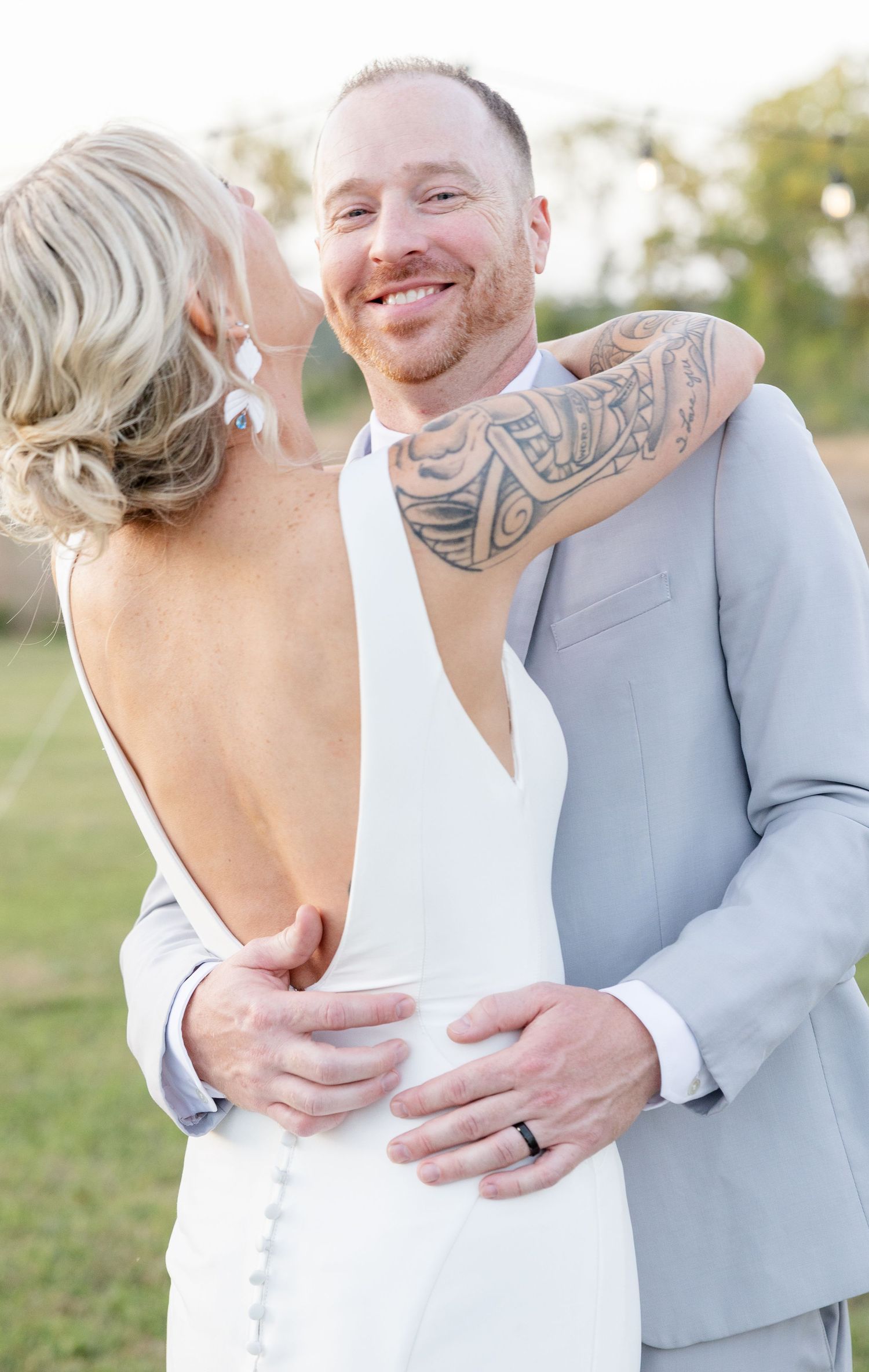 Smiling tattooed groom in gray suit embraces bride in backless white wedding gown outdoors.