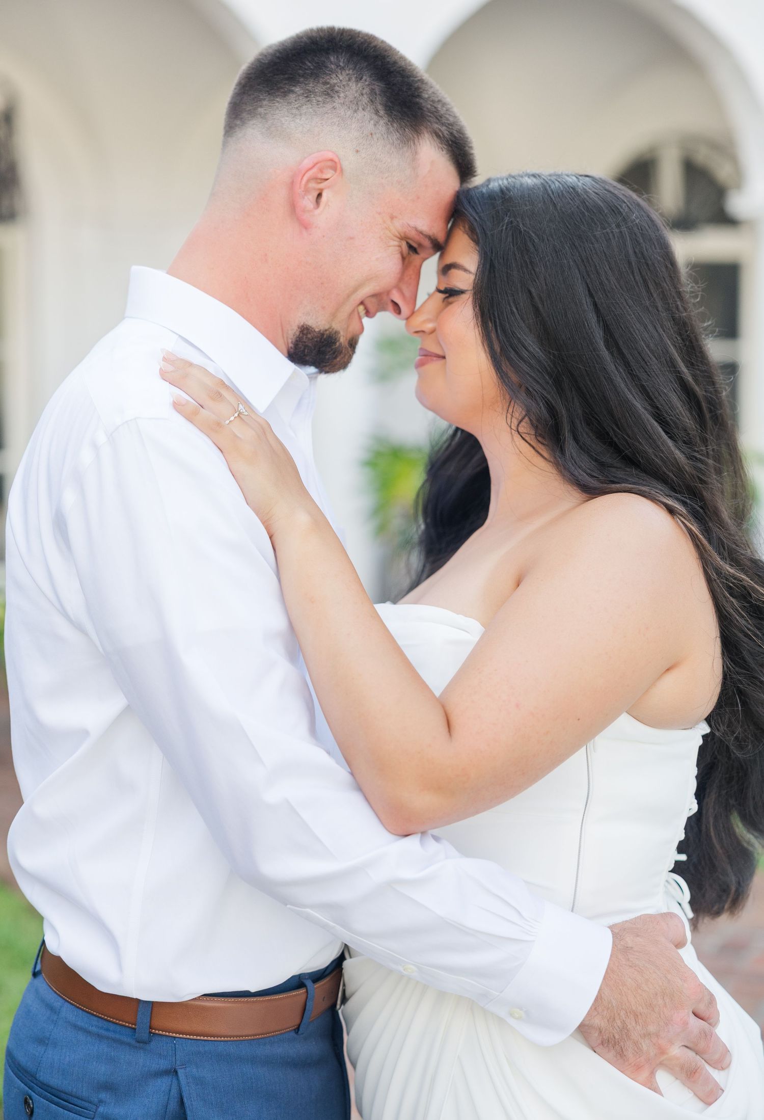 Engaged couple in white attire touching foreheads romantically in front of arched white building outdoors.