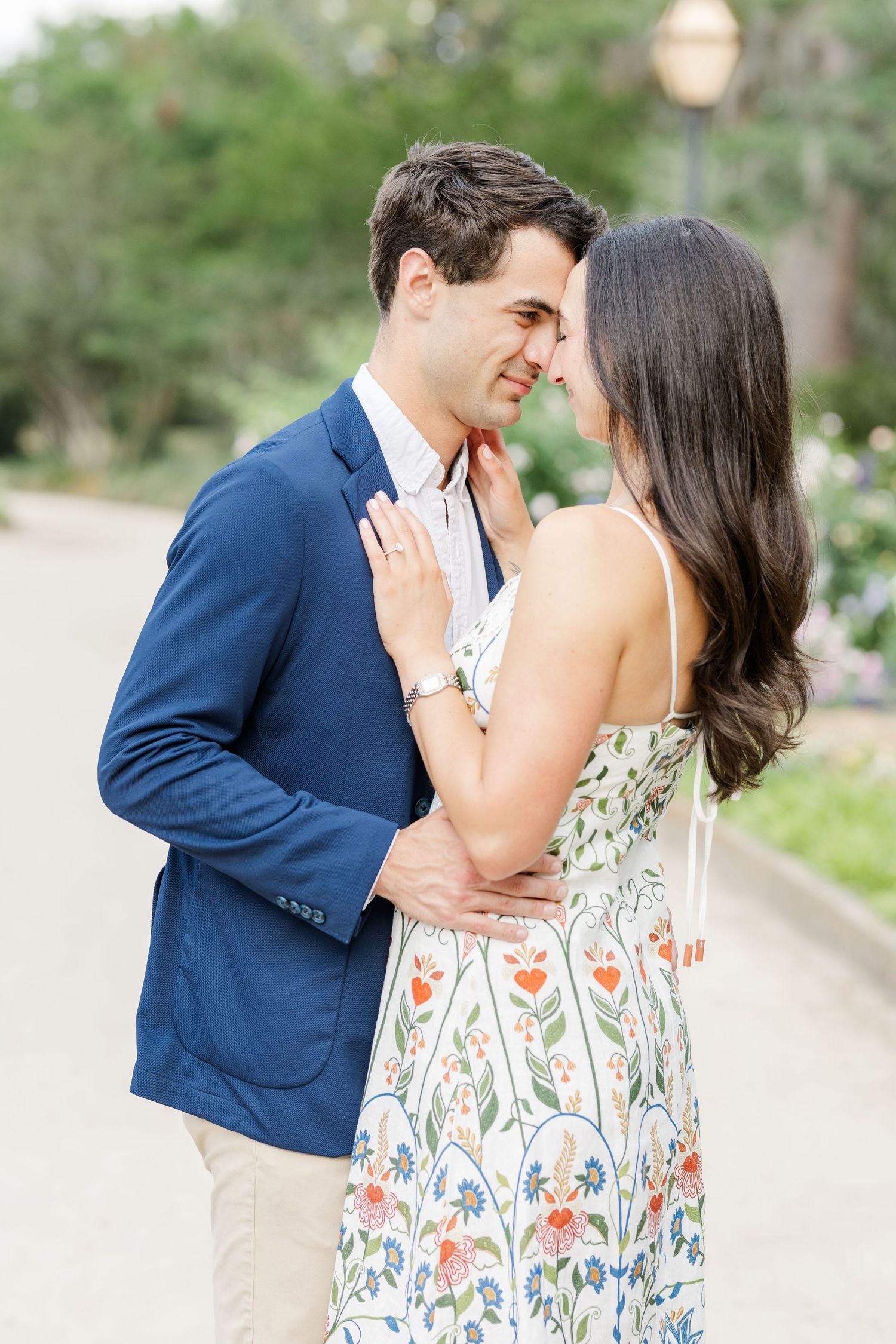 A couple in a romantic embrace, man in navy blazer and woman in floral dress, sharing an intimate moment outdoors.