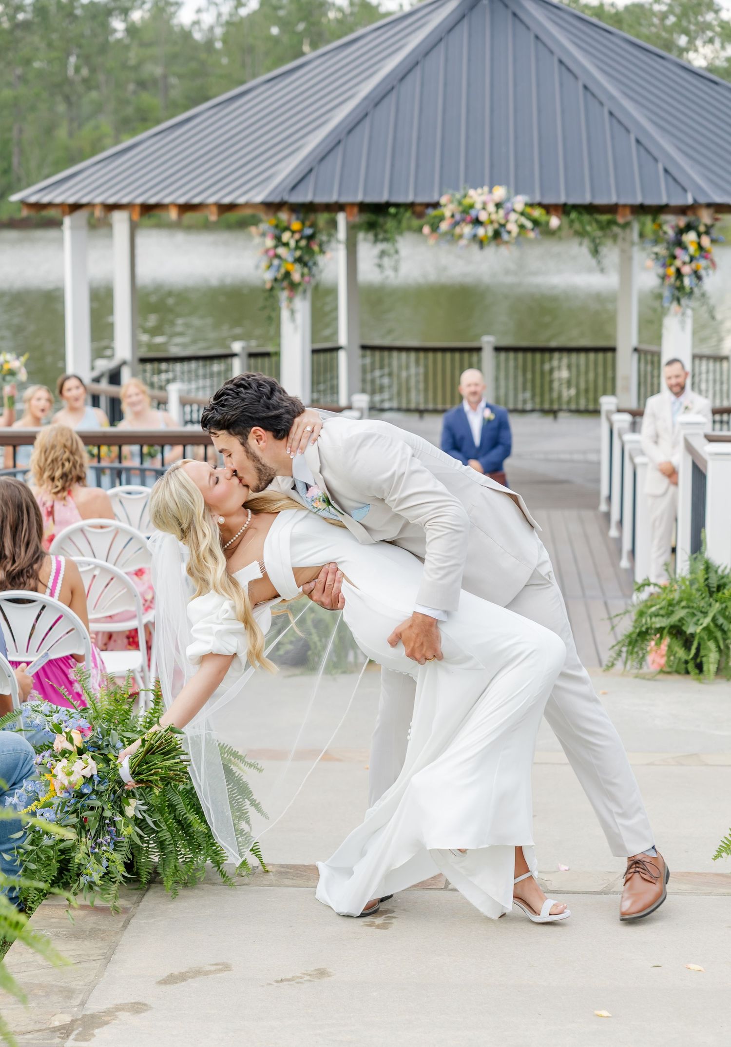 Groom dips bride for romantic kiss on dock walkway after outdoor lakeside wedding ceremony.