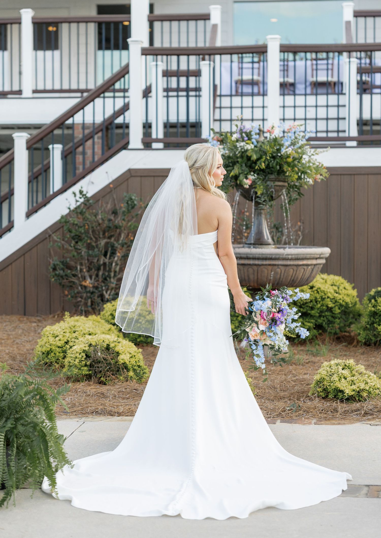 Back view of bride in fitted strapless white gown with cathedral veil holding colorful bouquet at venue.