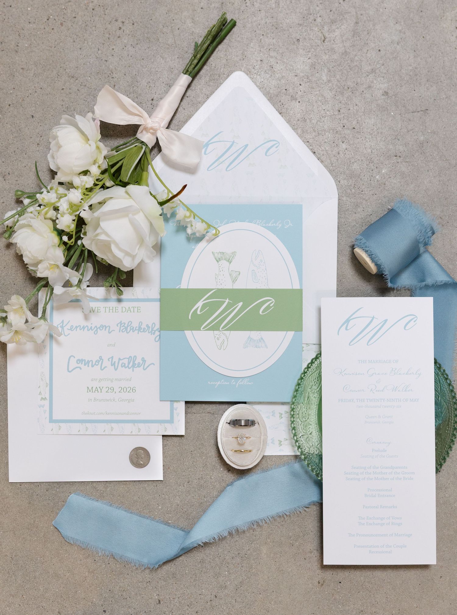 Elegant wedding invitation suite in blue and green with monogram, white roses, and silk ribbon on gray background.
