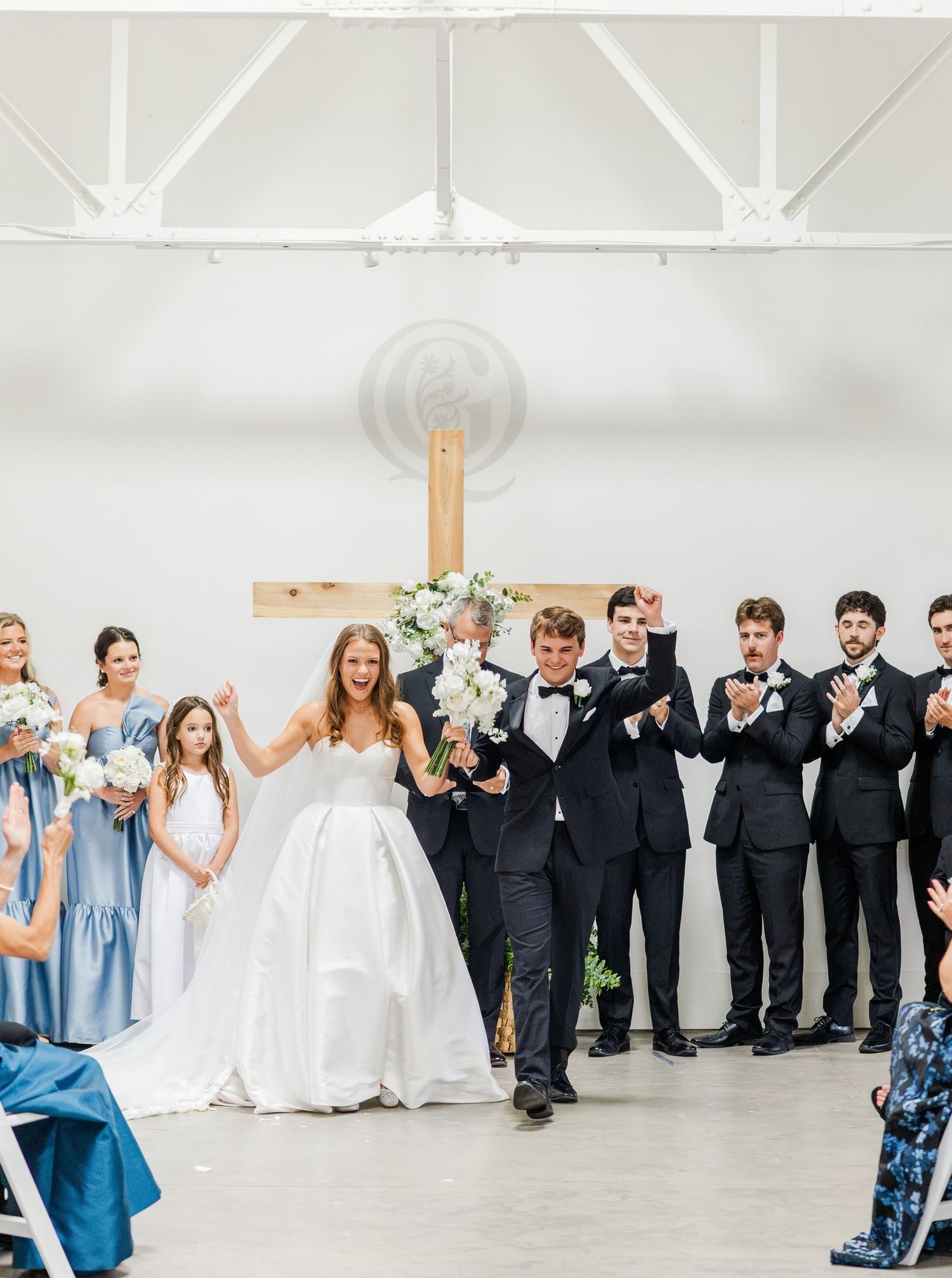 Joyful bride and groom celebrate with wedding party at altar featuring wooden cross in bright venue.