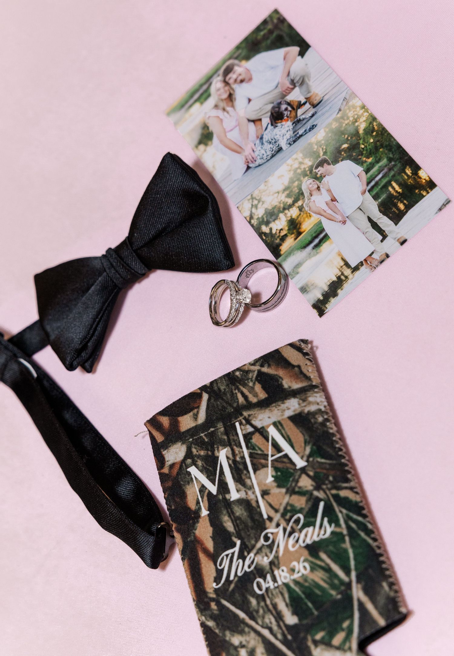 Wedding flat lay with black bow tie, gold rings, camo koozie, and couple photos on pink background.