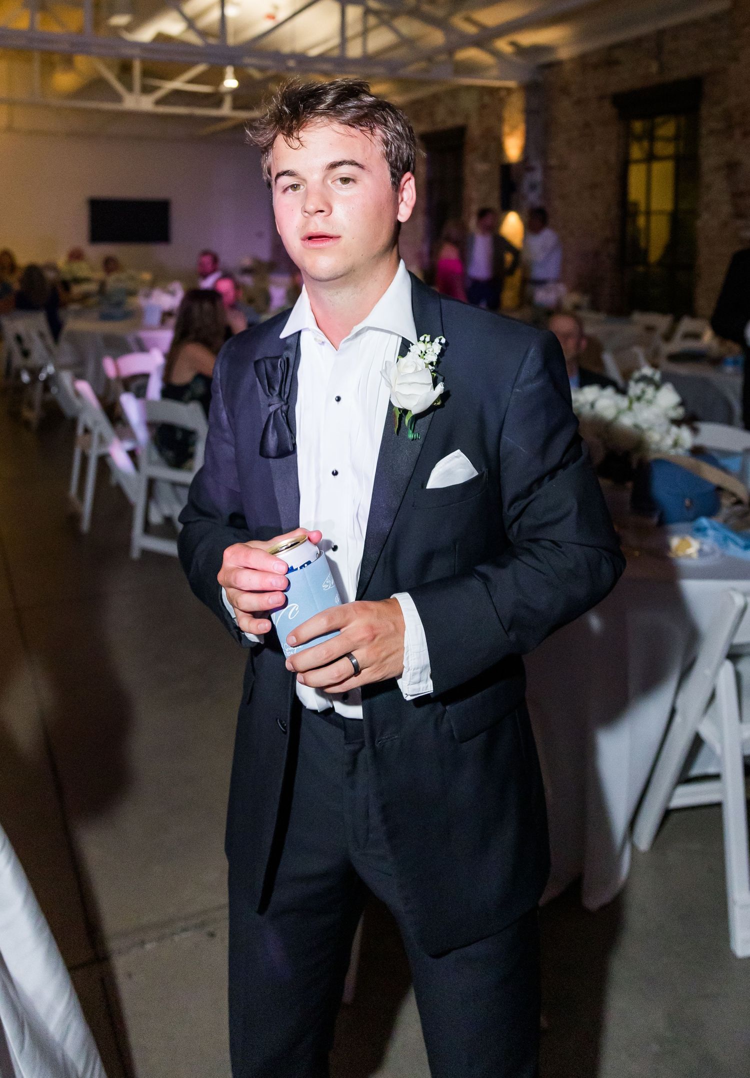 Young groomsman in black tuxedo holds a drink while standing in a warmly lit wedding reception hall.