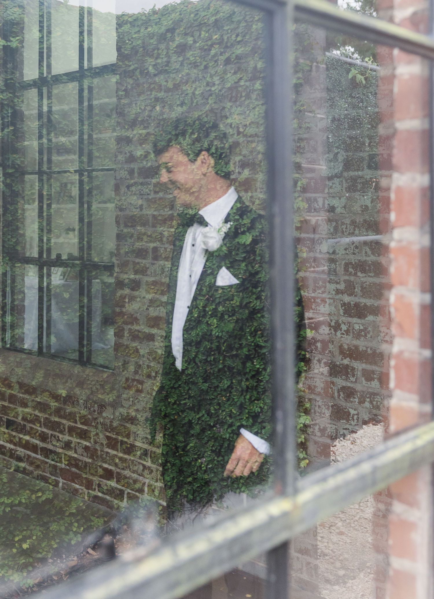 Groom in black tuxedo with white bow tie and boutonniere, photographed through glass window with ivy-covered brick wall.