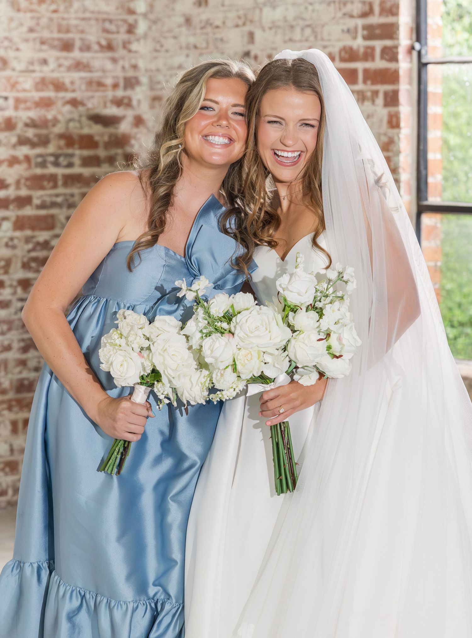 Smiling bride in white gown with veil and bridesmaid in dusty blue dress holding white bouquets.