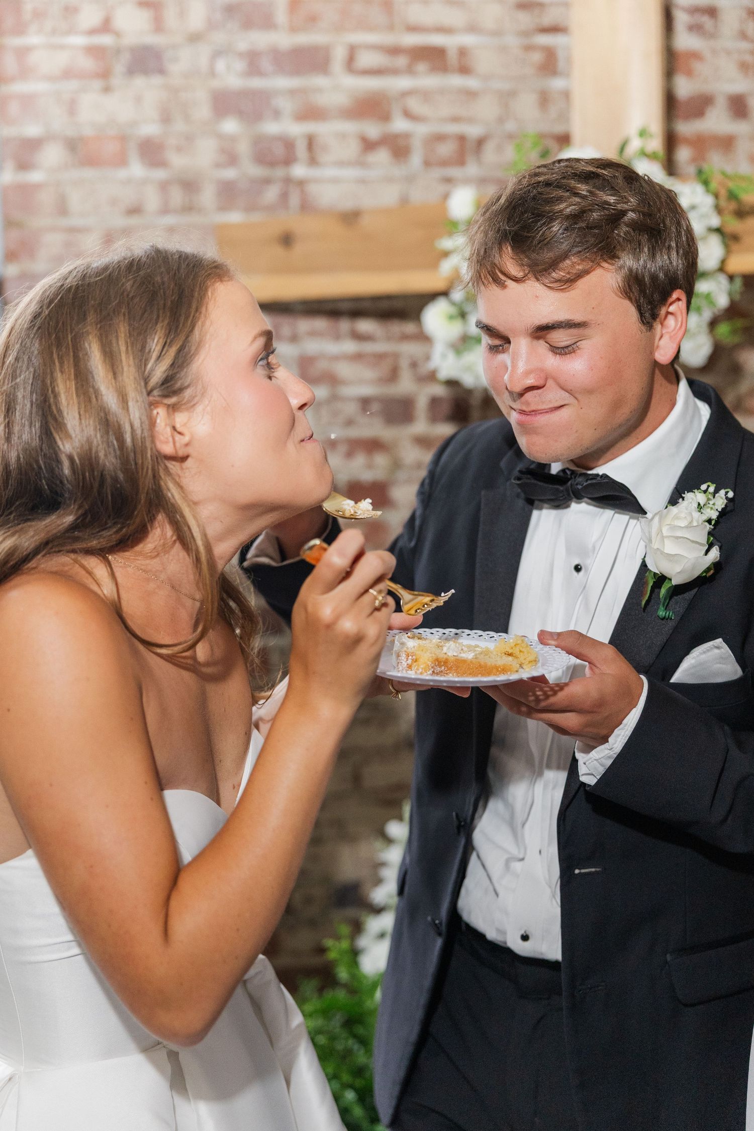 Bride and groom share a playful cake cutting moment at their wedding reception near a brick wall.