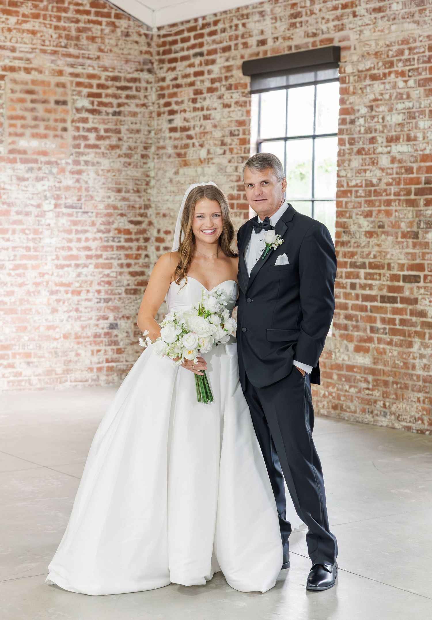 Bride and father pose together in brick-walled wedding venue with white bouquet.