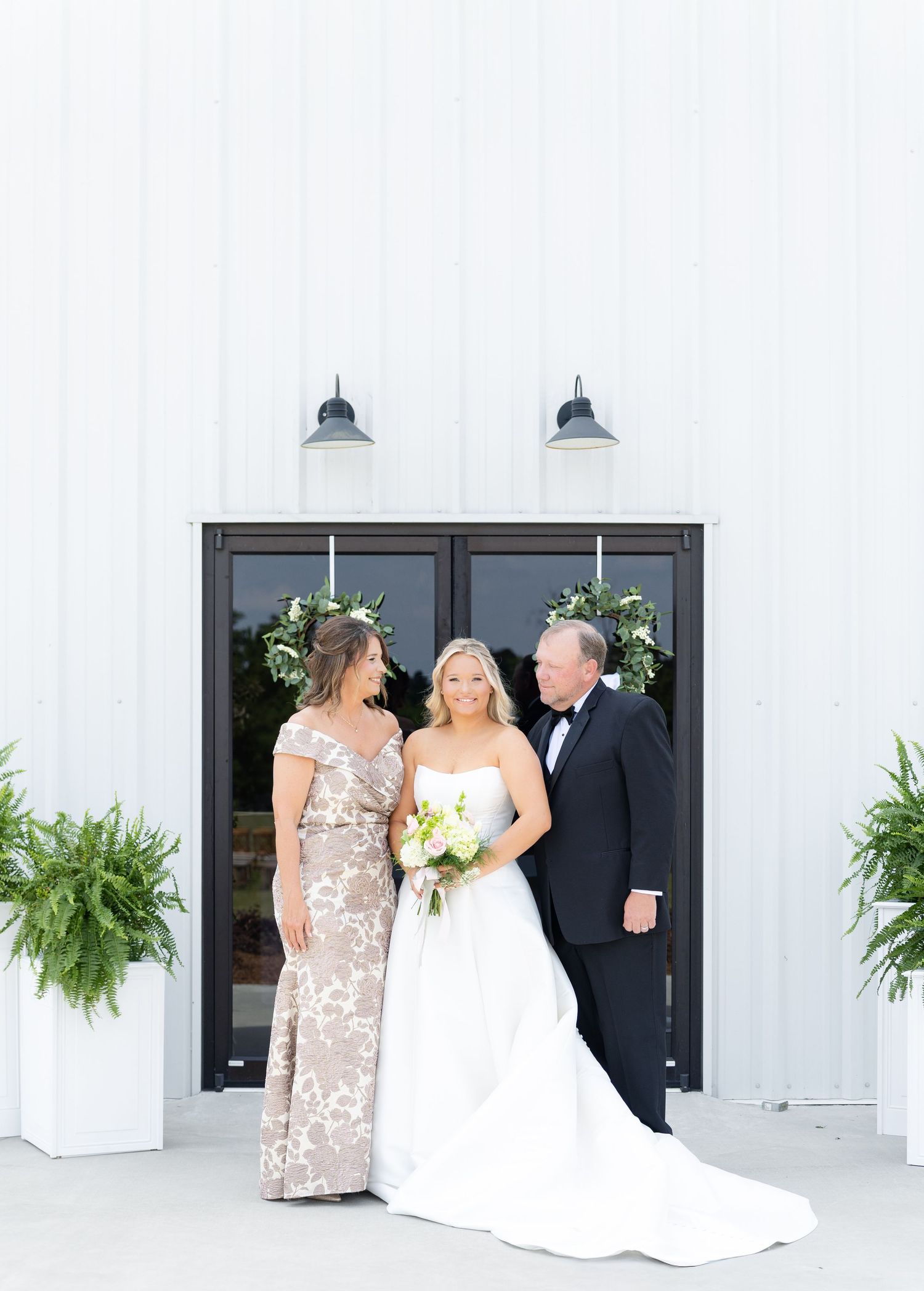 Bride with parents in front of white barn wedding venue doors decorated with greenery wreaths.