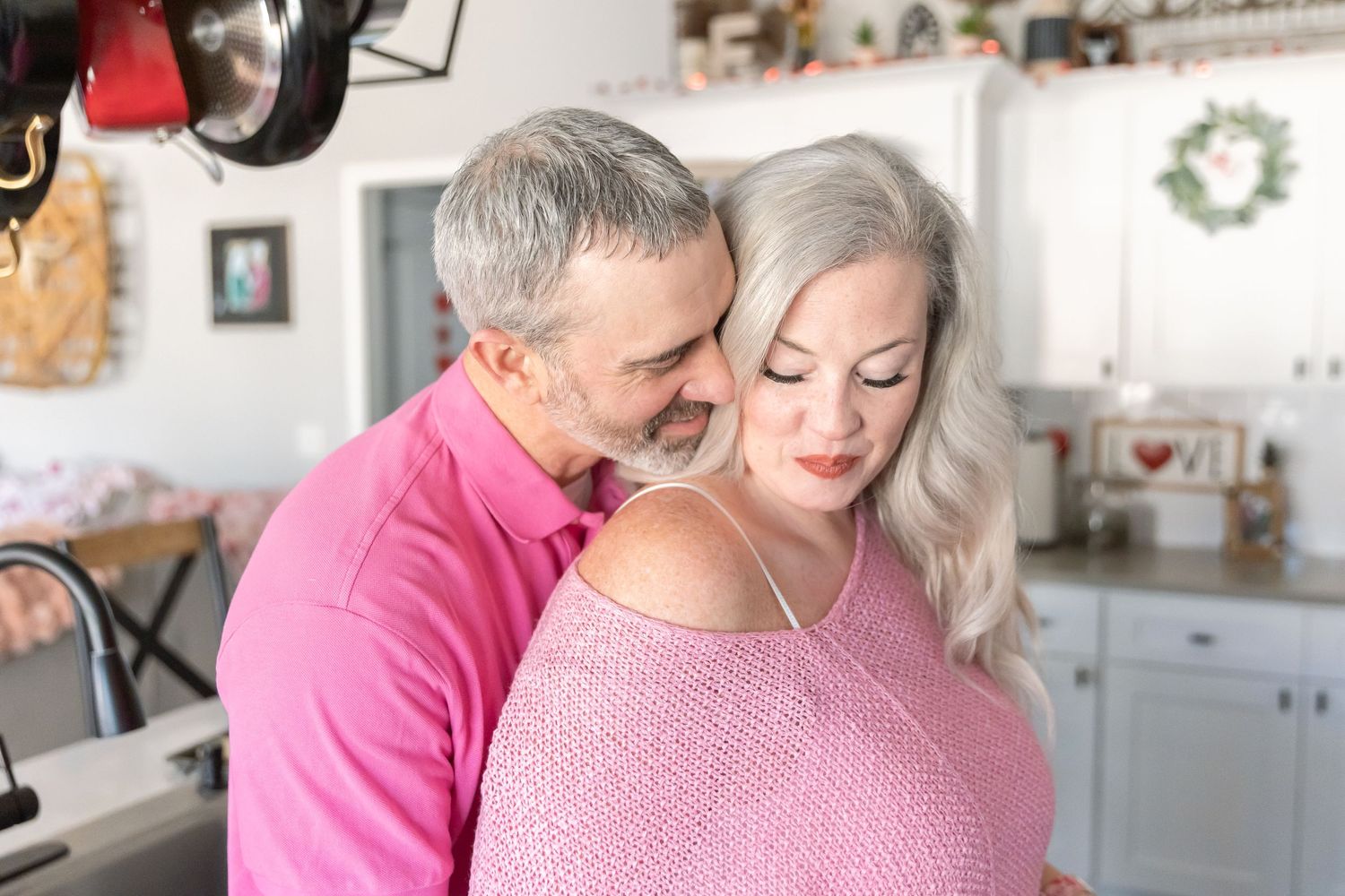 A couple embraces in their kitchen wearing pink and white sweaters while sharing an intimate moment.