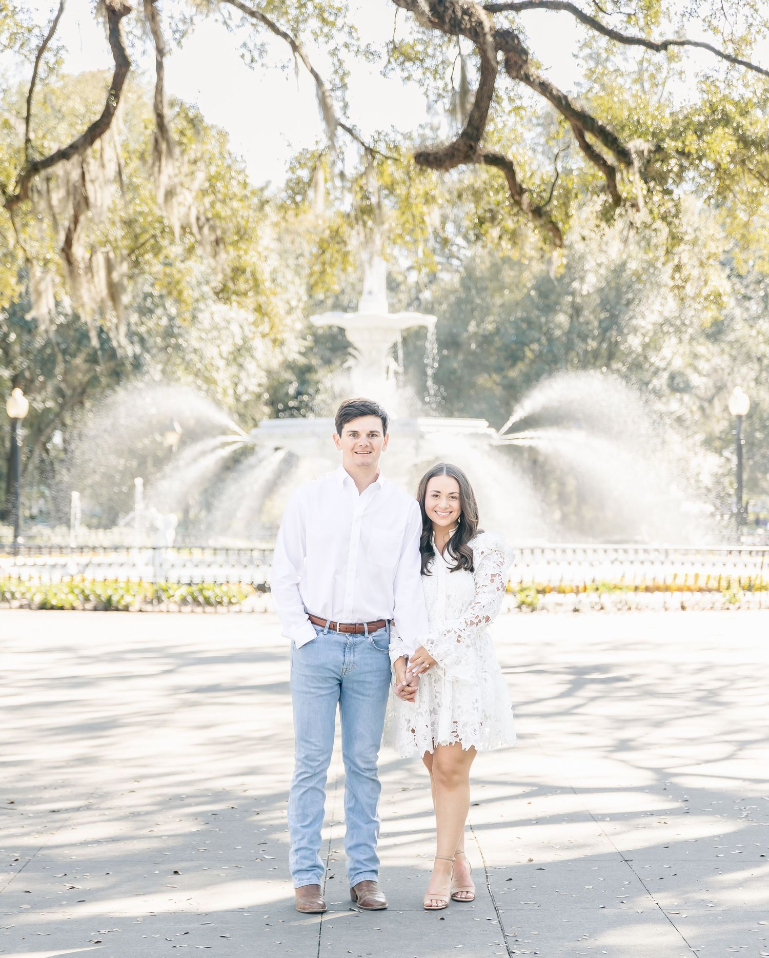 A couple stands together in front of an ornate fountain with Spanish moss hanging from trees above.