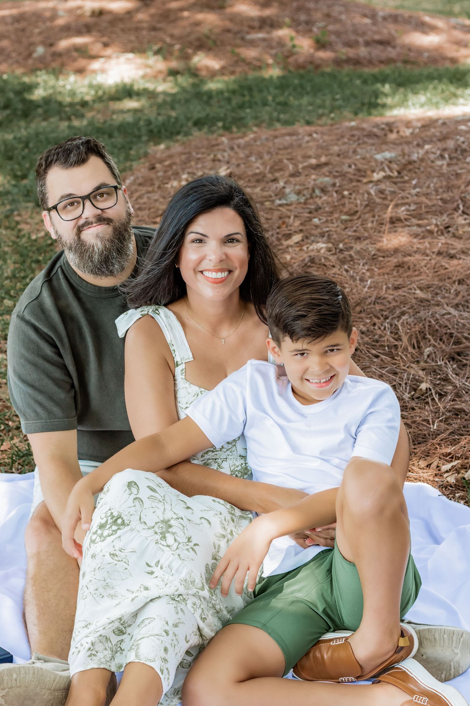 A family of three poses for a casual outdoor portrait while sitting on a blanket.