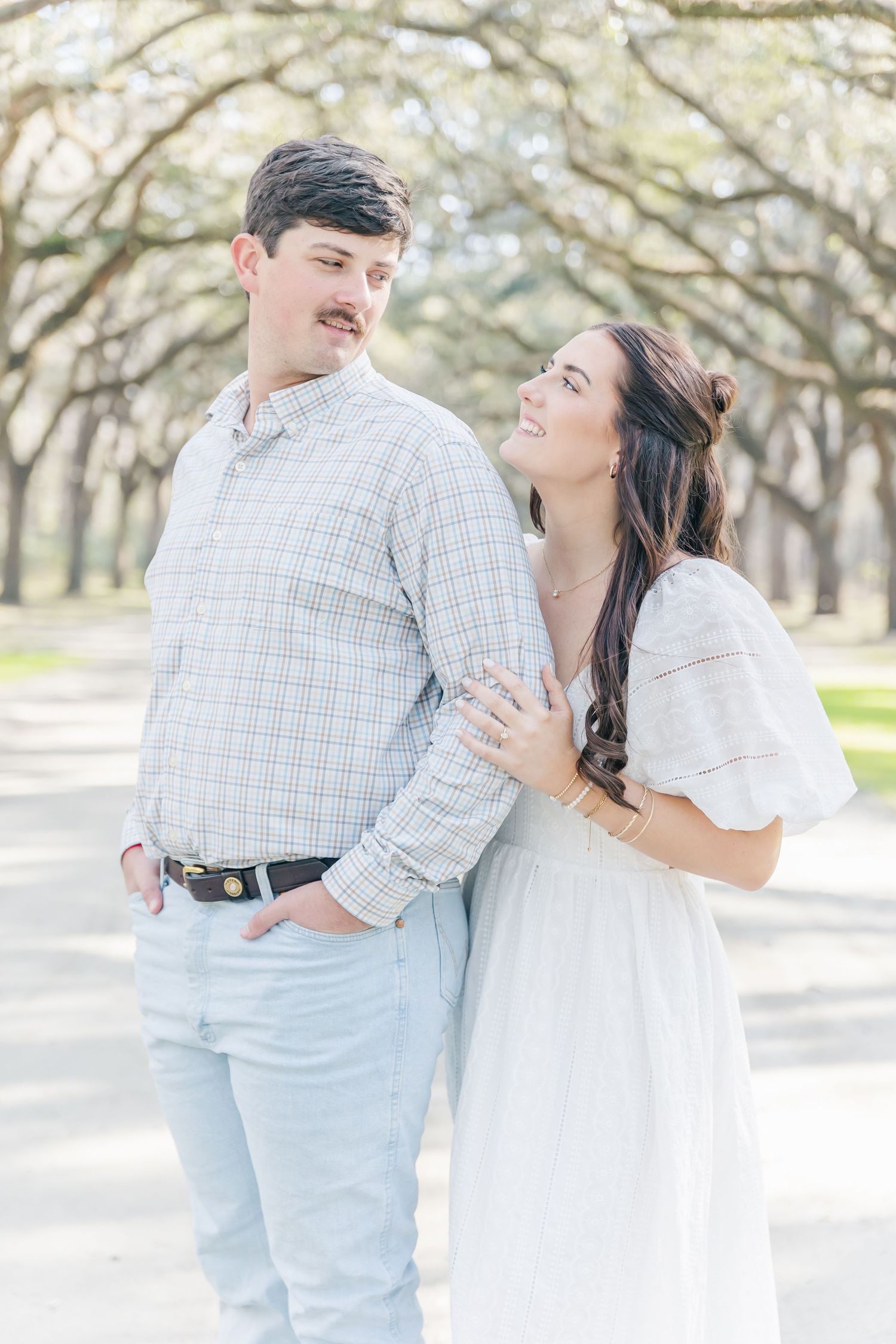 A couple shares a candid laugh while posing under a canopy of oak trees.