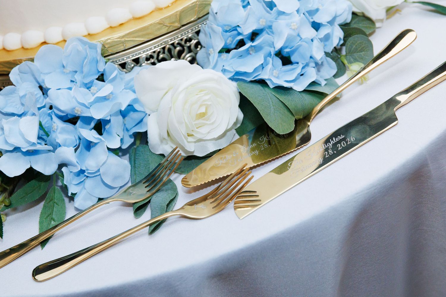 Gold cake cutting set with blue hydrangeas and white roses displayed on a wedding reception table.