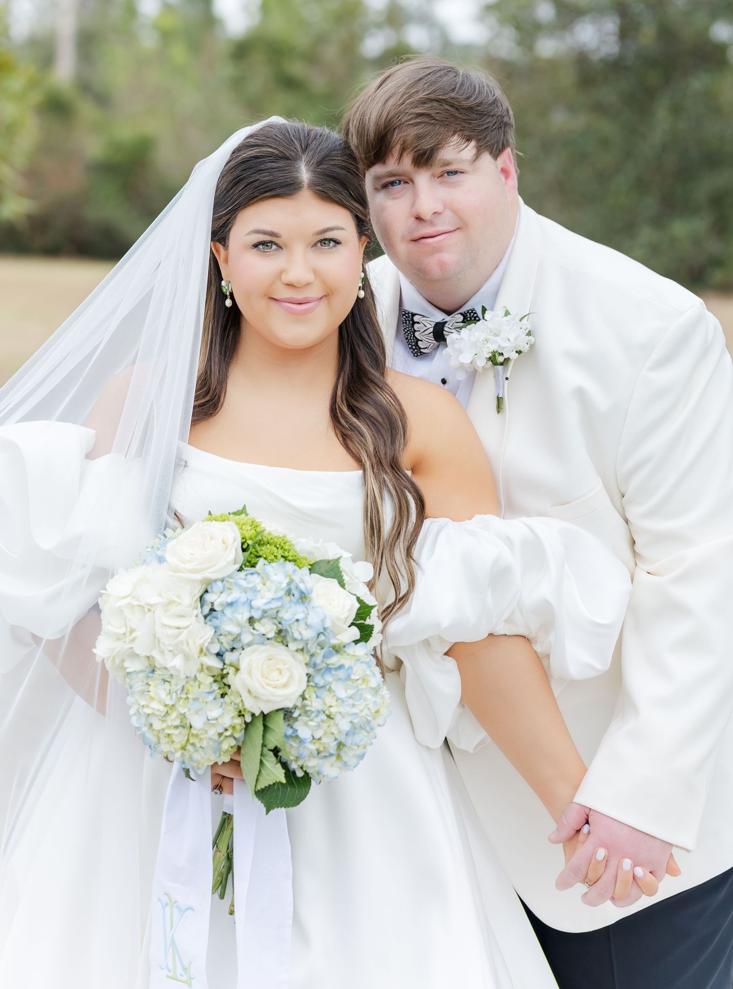 Bride in white gown holding blue and white bouquet poses with groom in white tuxedo outdoors.