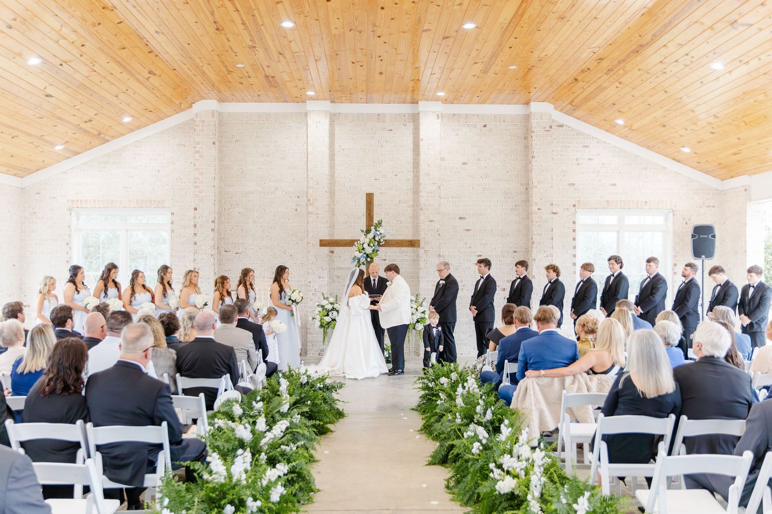 Wedding ceremony inside a white chapel with wooden ceiling, cross altar, and full bridal party.