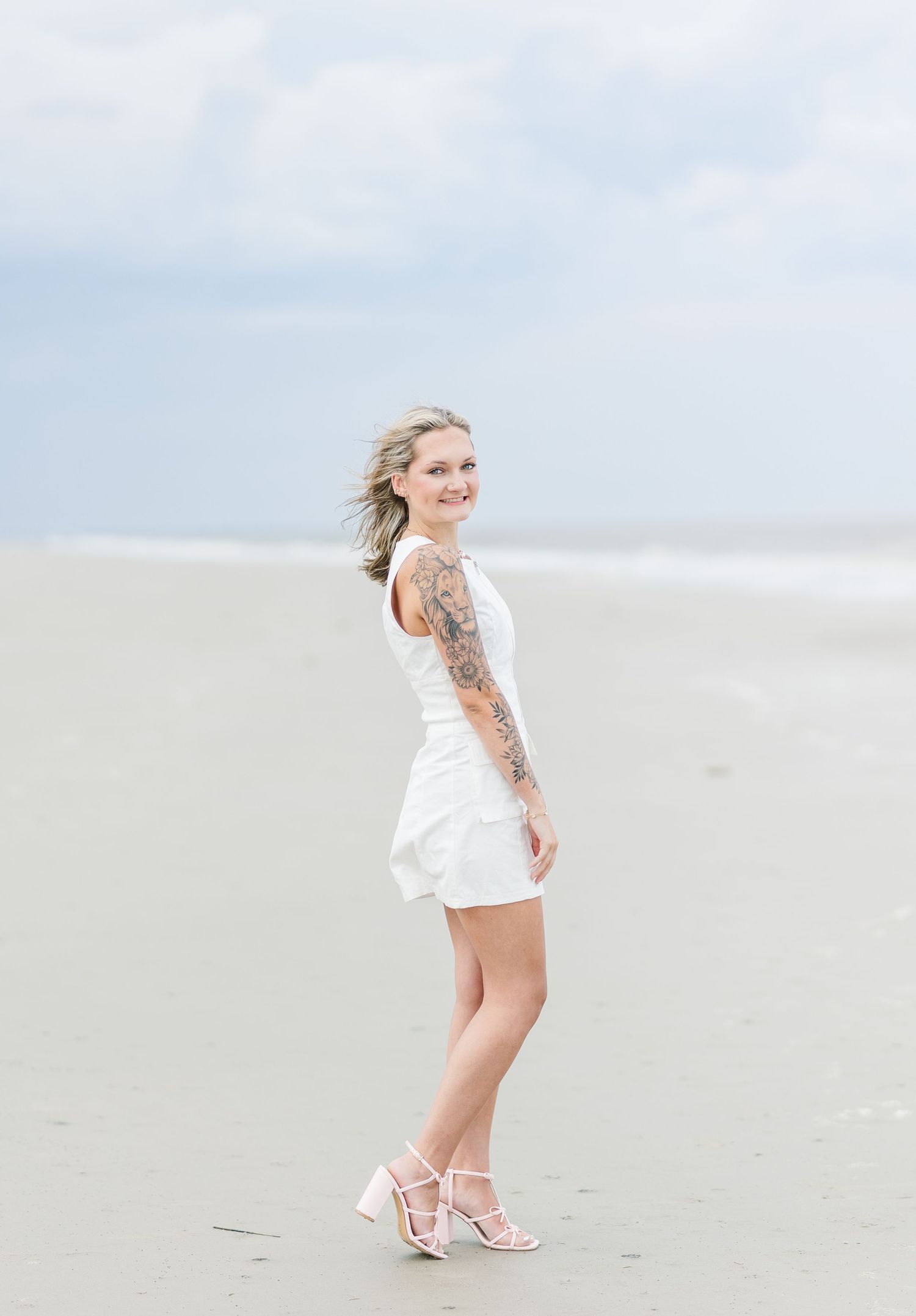 Graduate in white dress poses on beach with gentle waves in background.