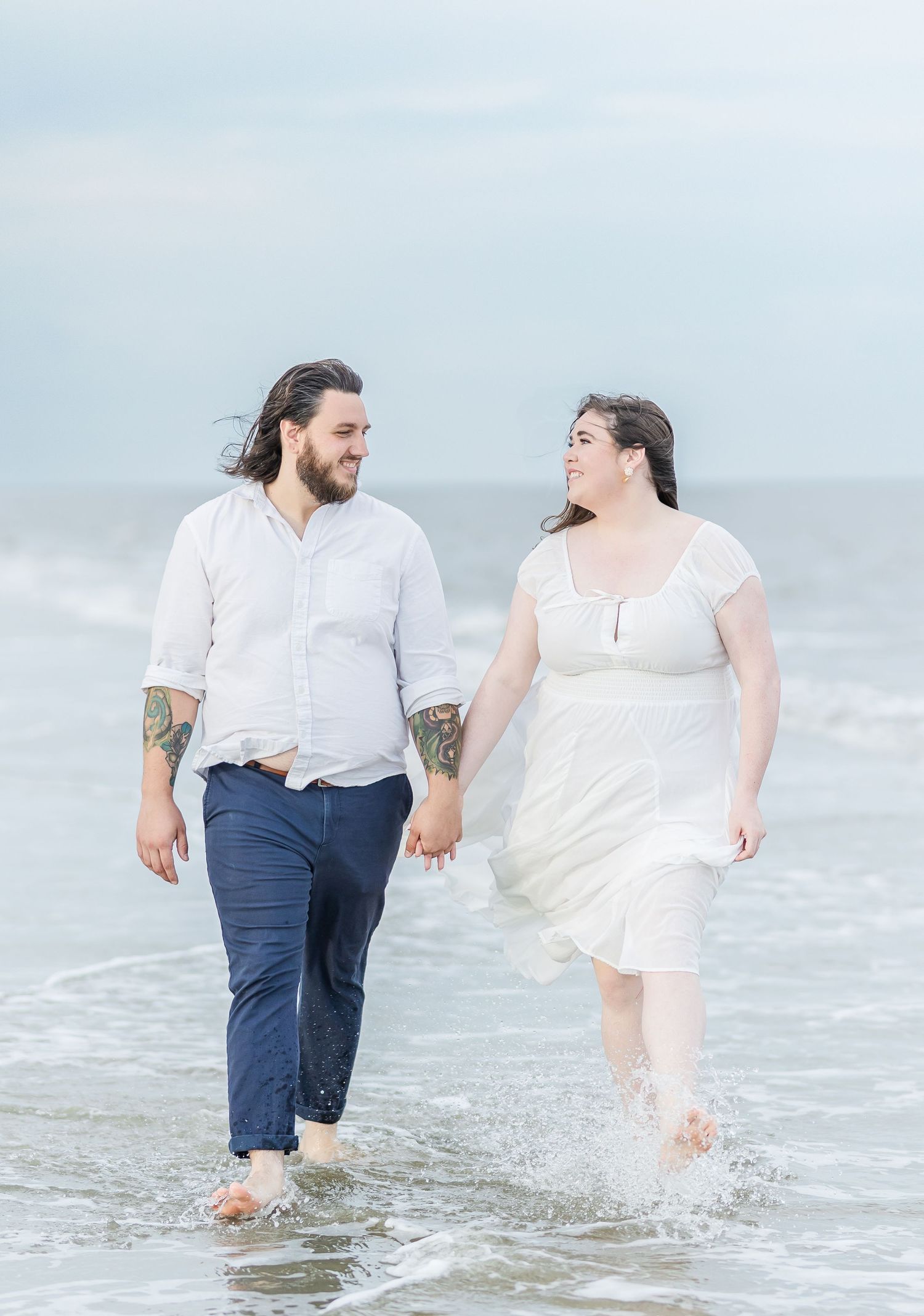 A couple walks through shallow ocean waves while holding hands.
