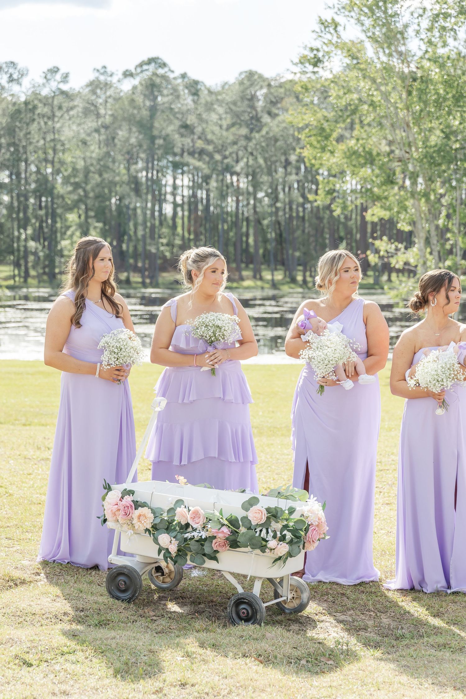 Bridesmaids in lilac dresses stand with bouquets next to decorative wagon in outdoor setting.