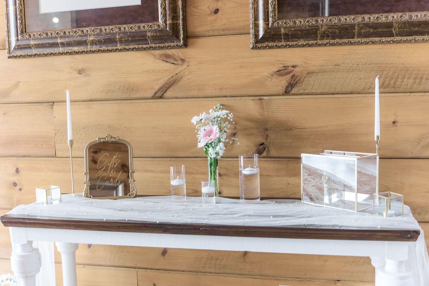Wedding reception table setup with white candles and frames against wooden wall decor.