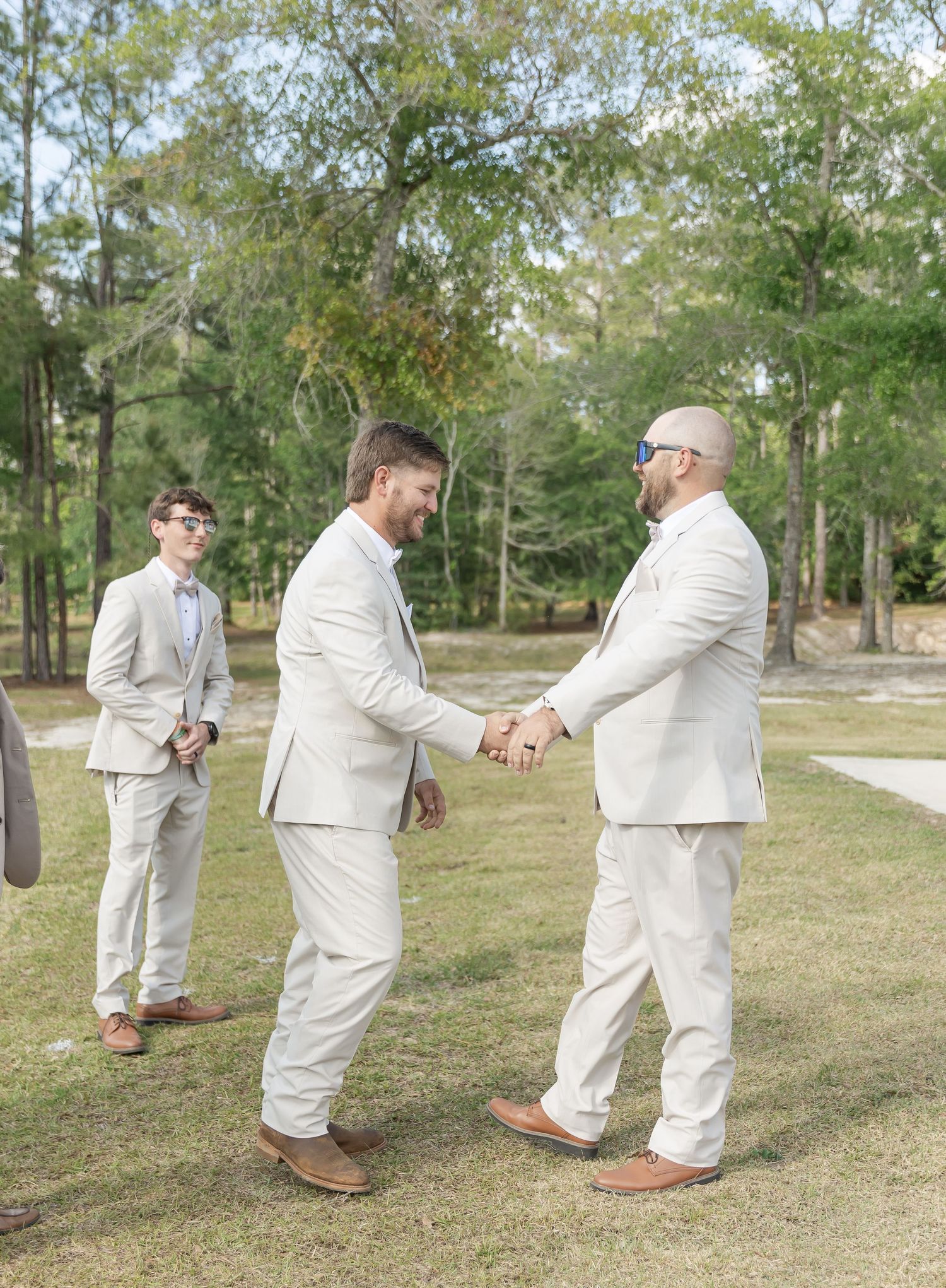 Groomsmen in light beige suits shake hands during an outdoor wedding ceremony.
