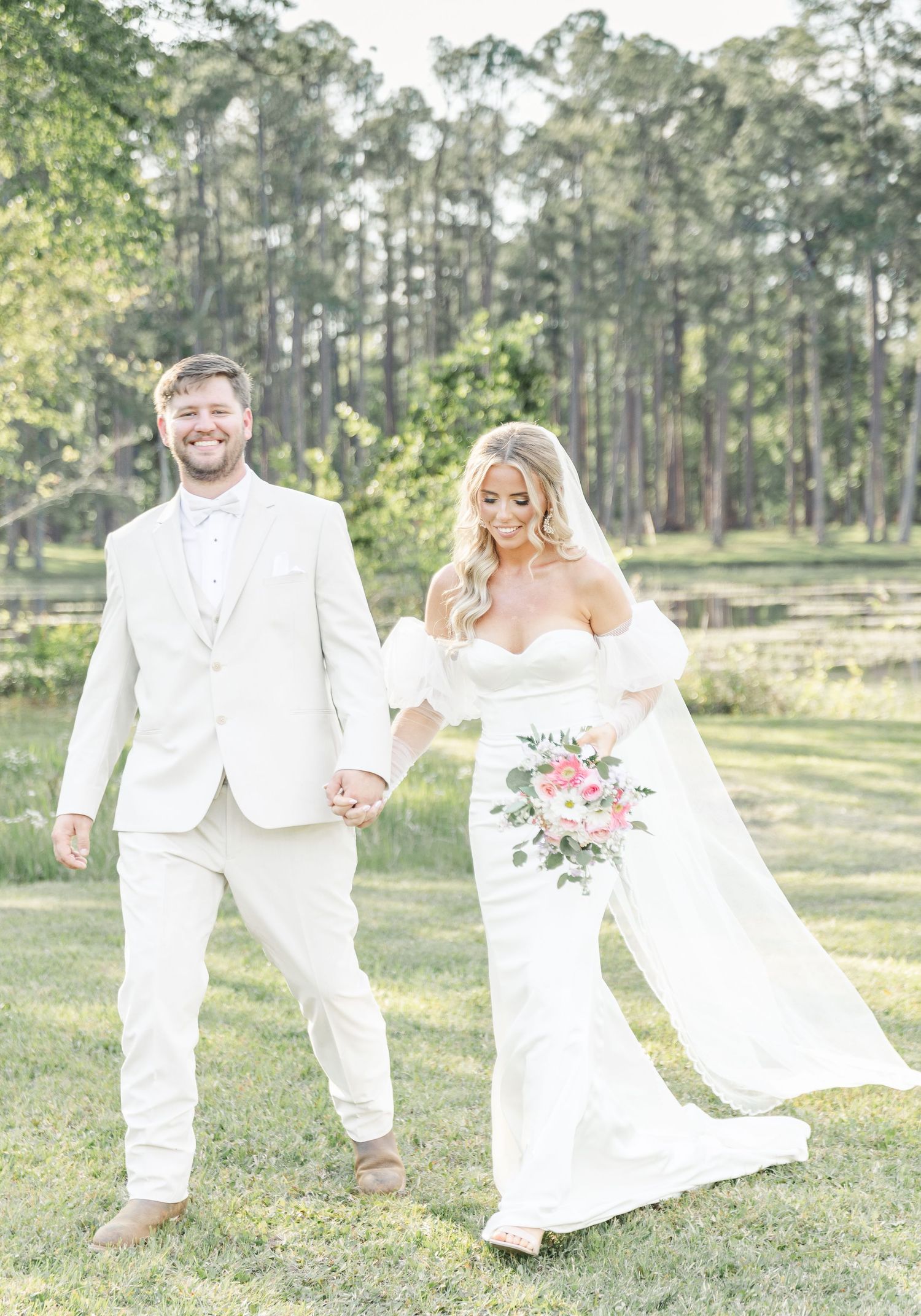 Newlyweds in all-white attire walk hand in hand through a scenic pine forest setting.